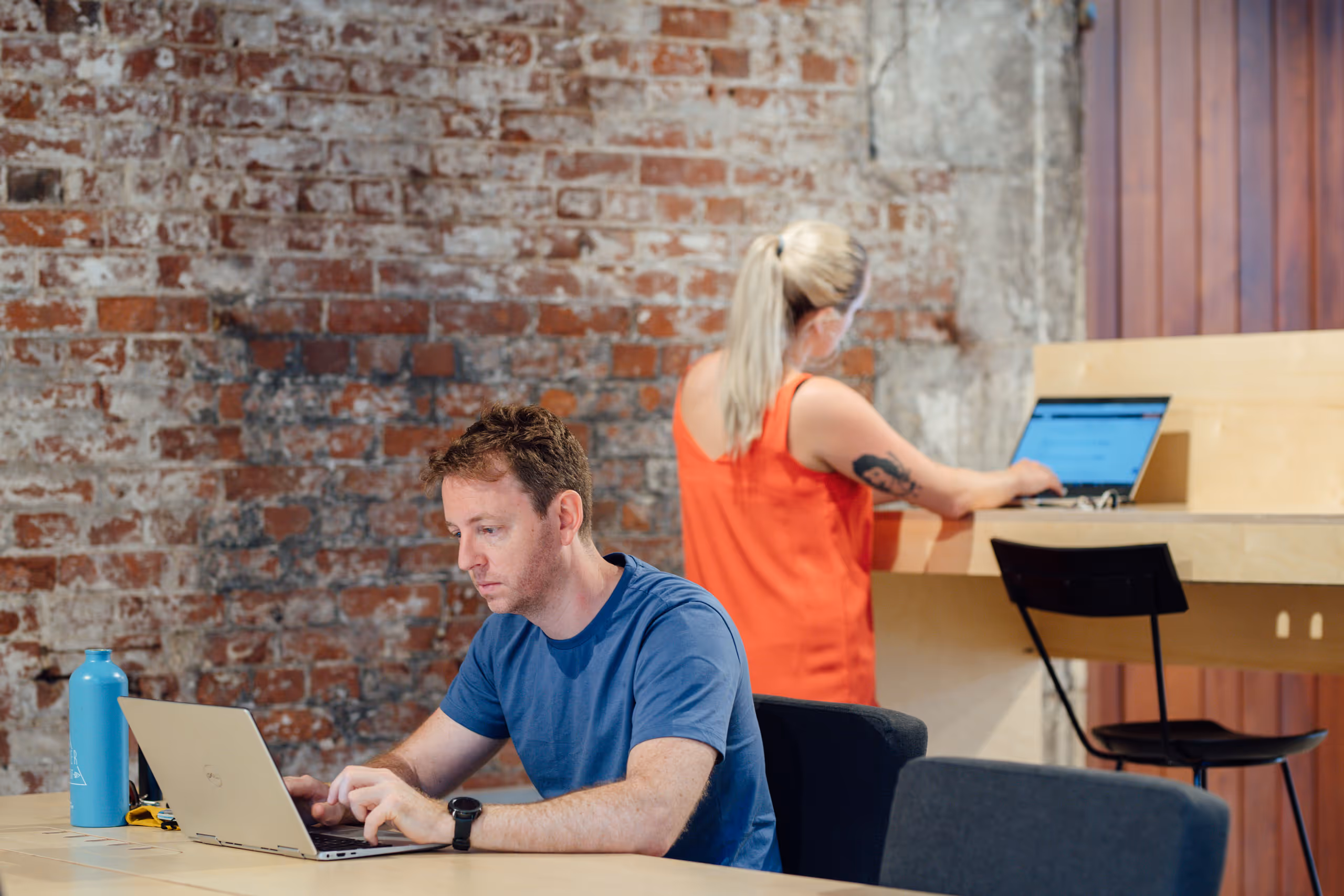 Two people working on laptops at wooden tables in industrial setting with weathered brick walls and modern furniture