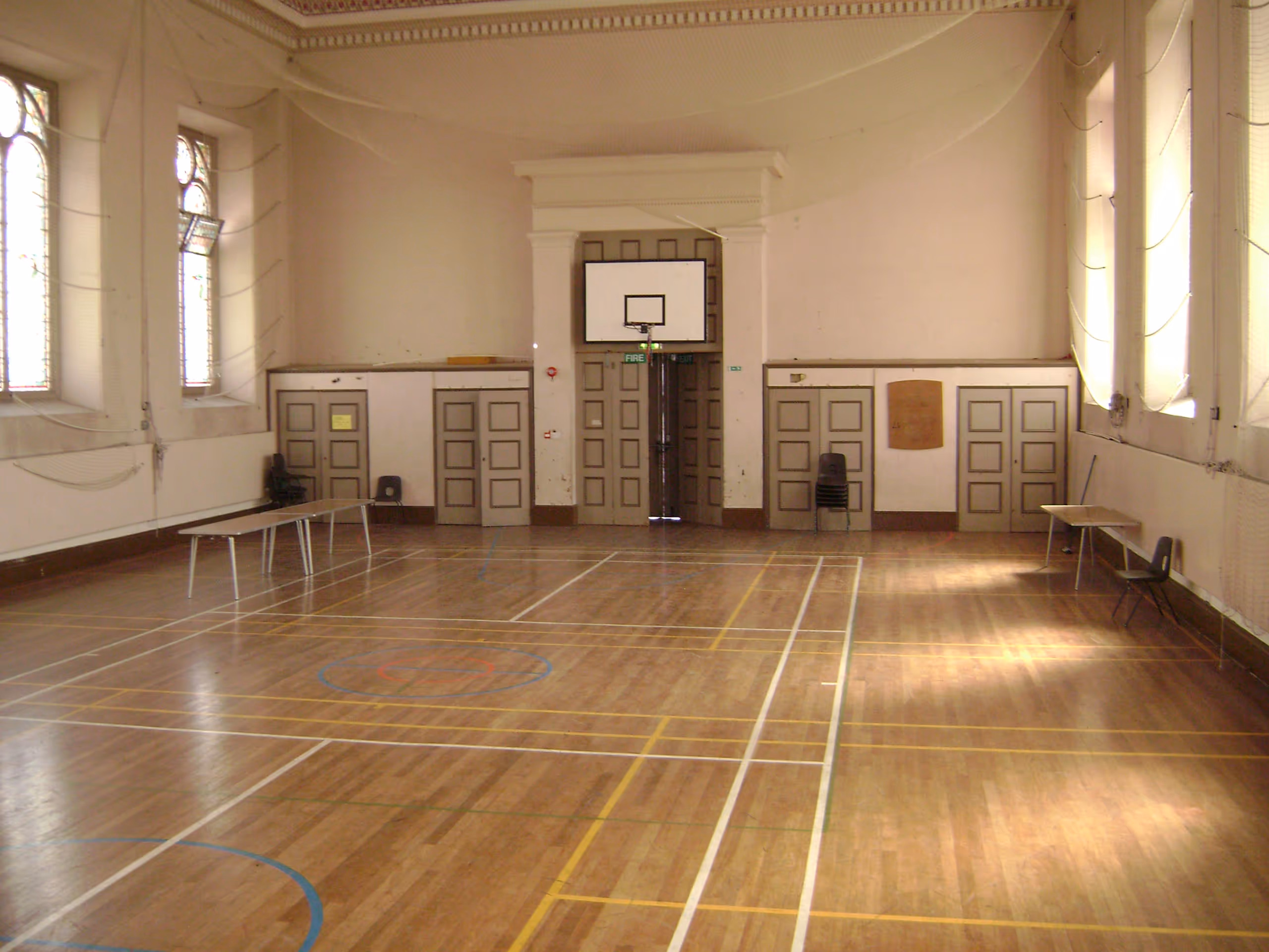 Historic hall interior with wooden floors, basketball court markings, ornate ceiling, and tall arched windows