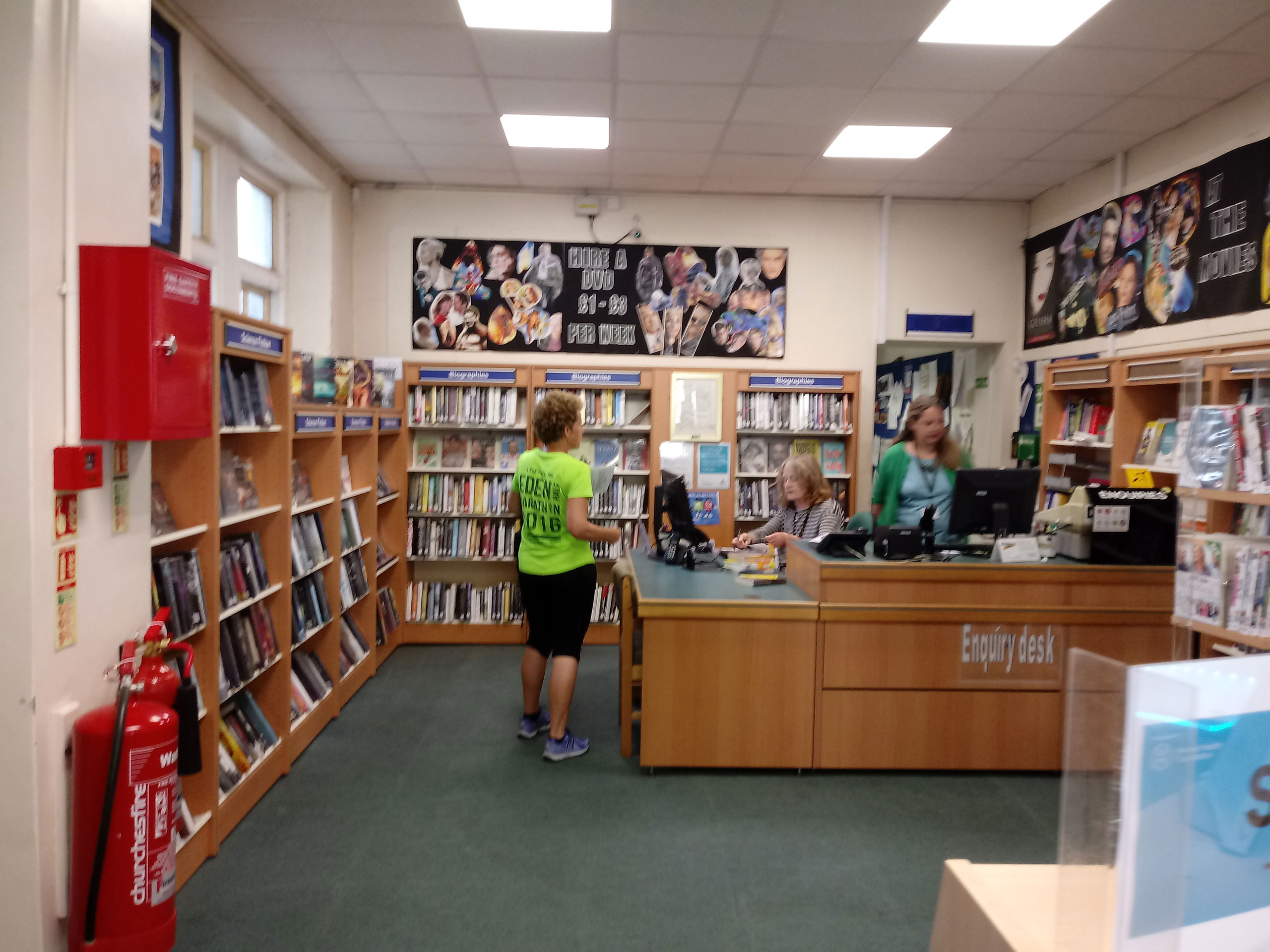 Modern library interior with wooden shelving, enquiry desk, and patrons browsing books and DVDs