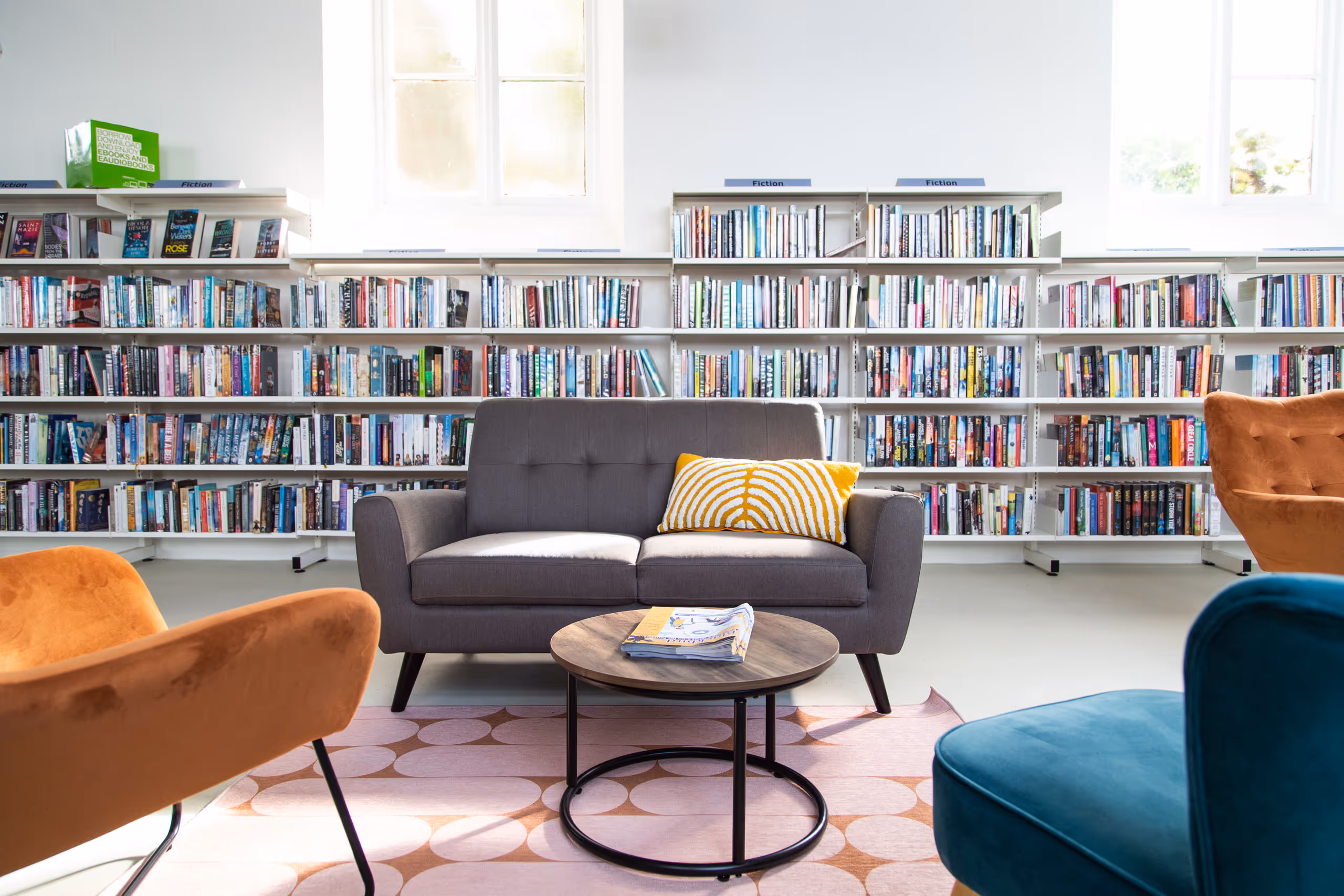 Comfortable reading lounge with gray sofa, orange velvet chairs, and extensive fiction collection on white shelves