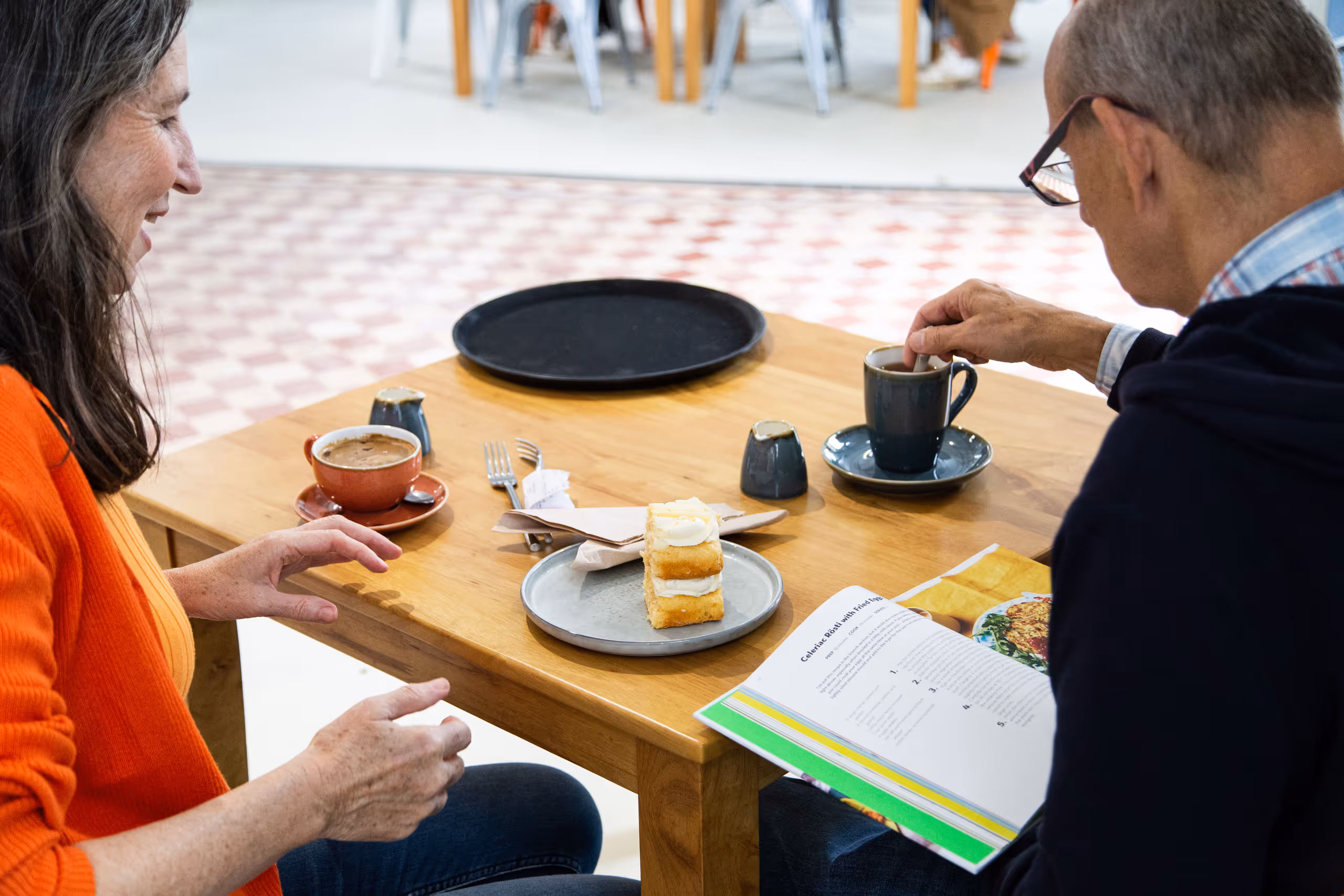 Two people enjoying coffee and cake at wooden table in library café area with geometric floor pattern