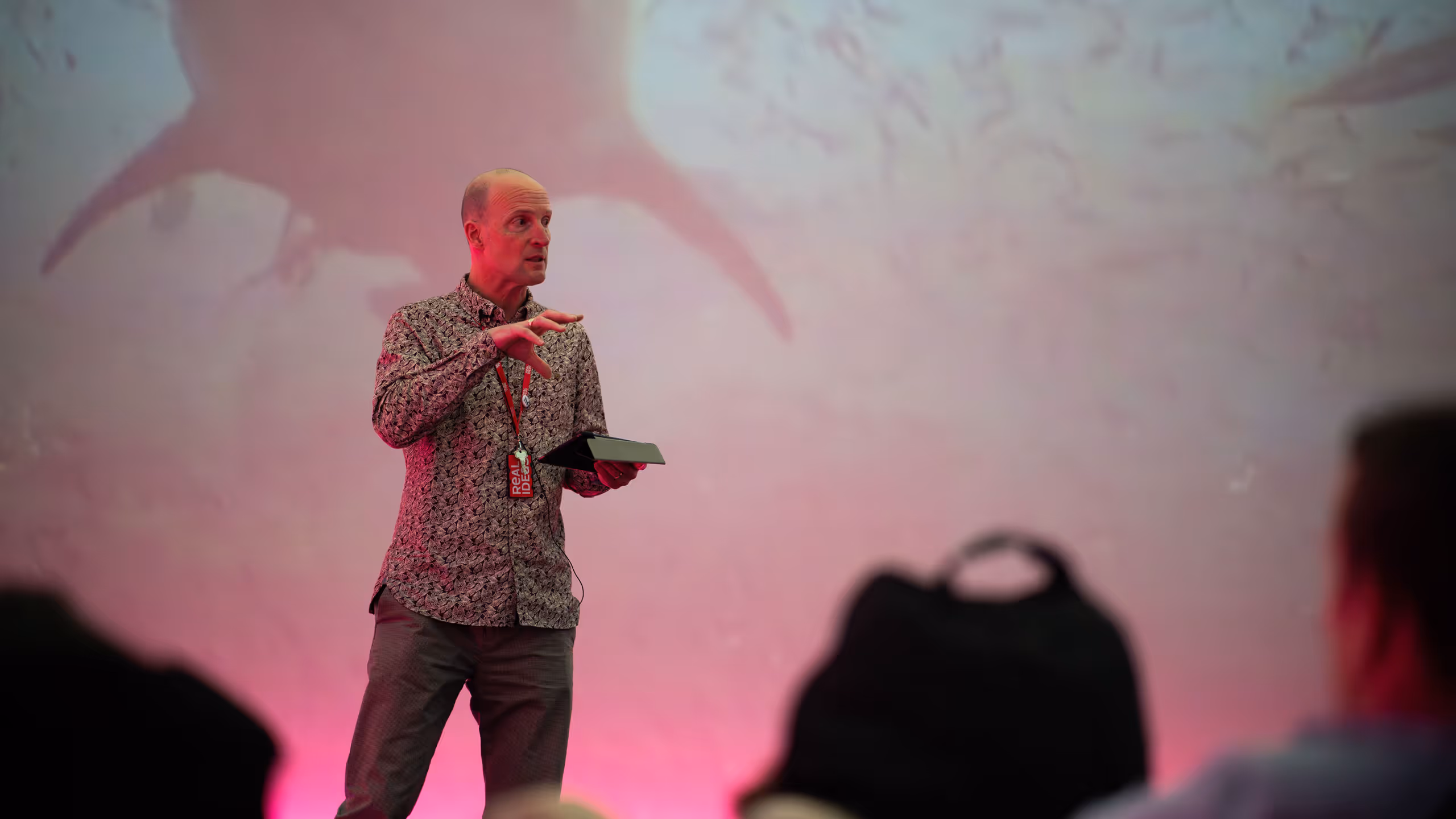 Speaker presenting to audience with pink lighting and projected visuals behind stage