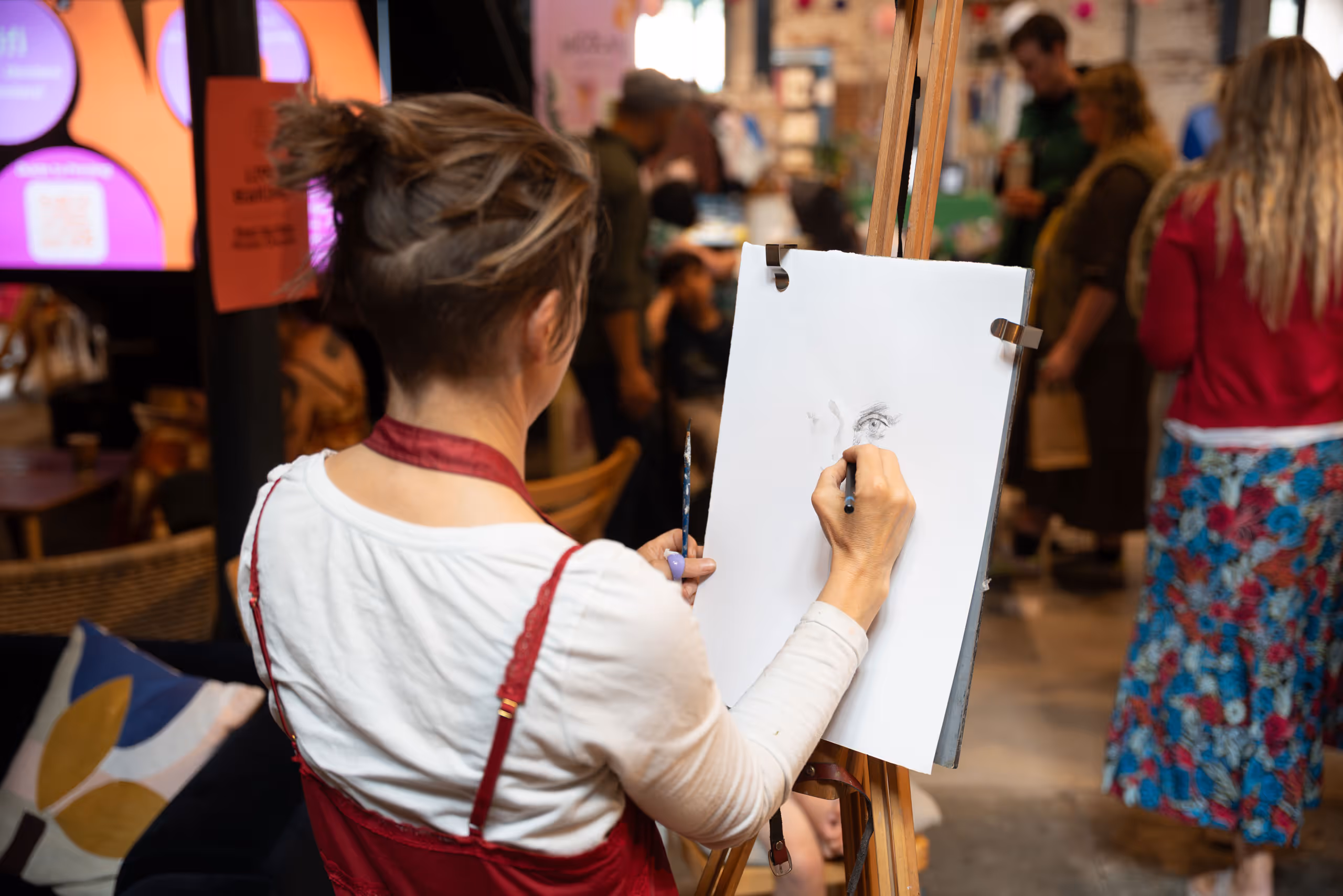 Artist drawing on easel during community art event with people socializing in background