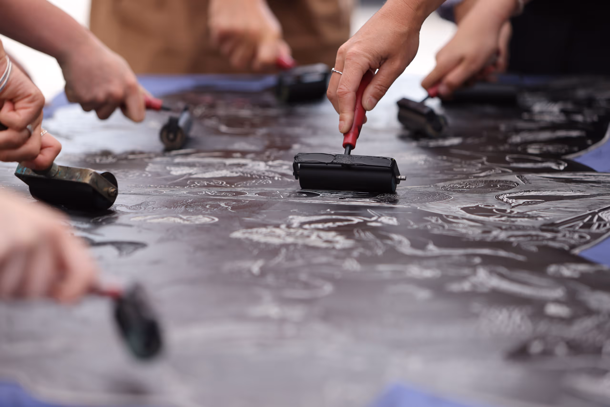 Multiple hands using ink rollers on large printing surface creating detailed artwork