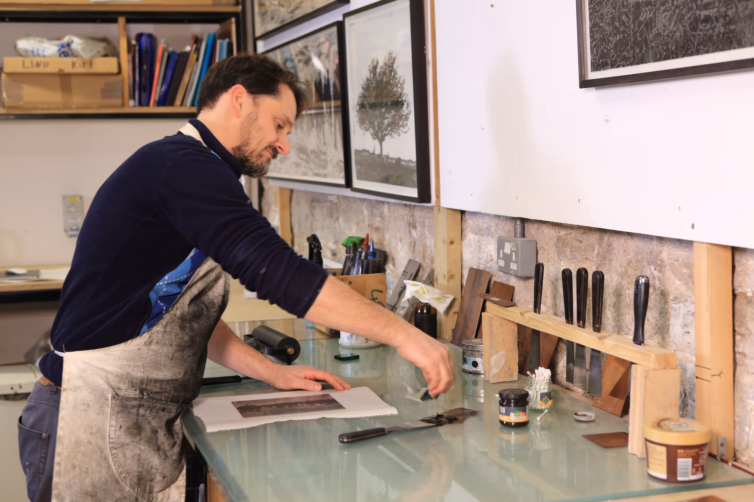 Artist in apron working at glass-topped table with printmaking tools and supplies in organized studio