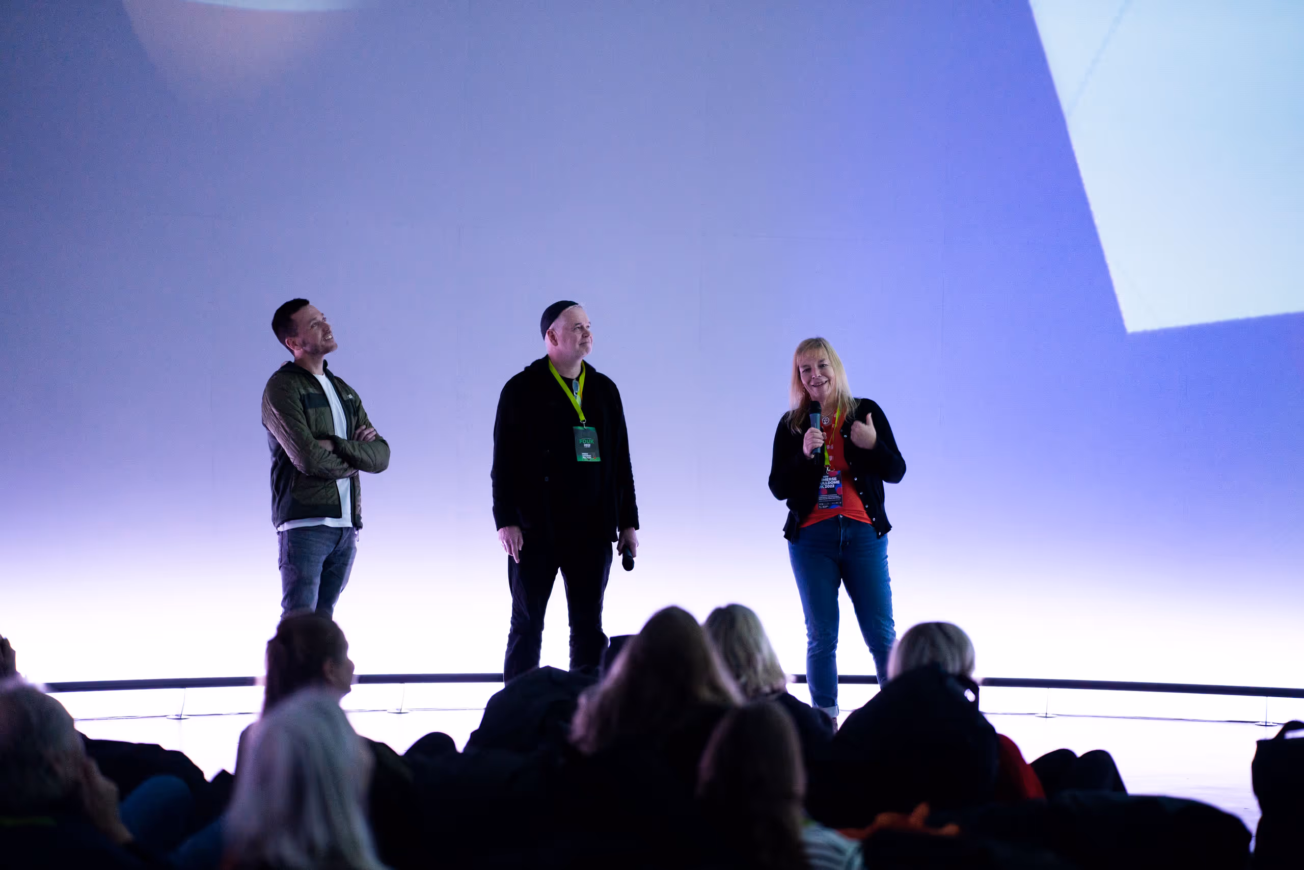 Three speakers presenting to seated audience on illuminated stage with purple lighting