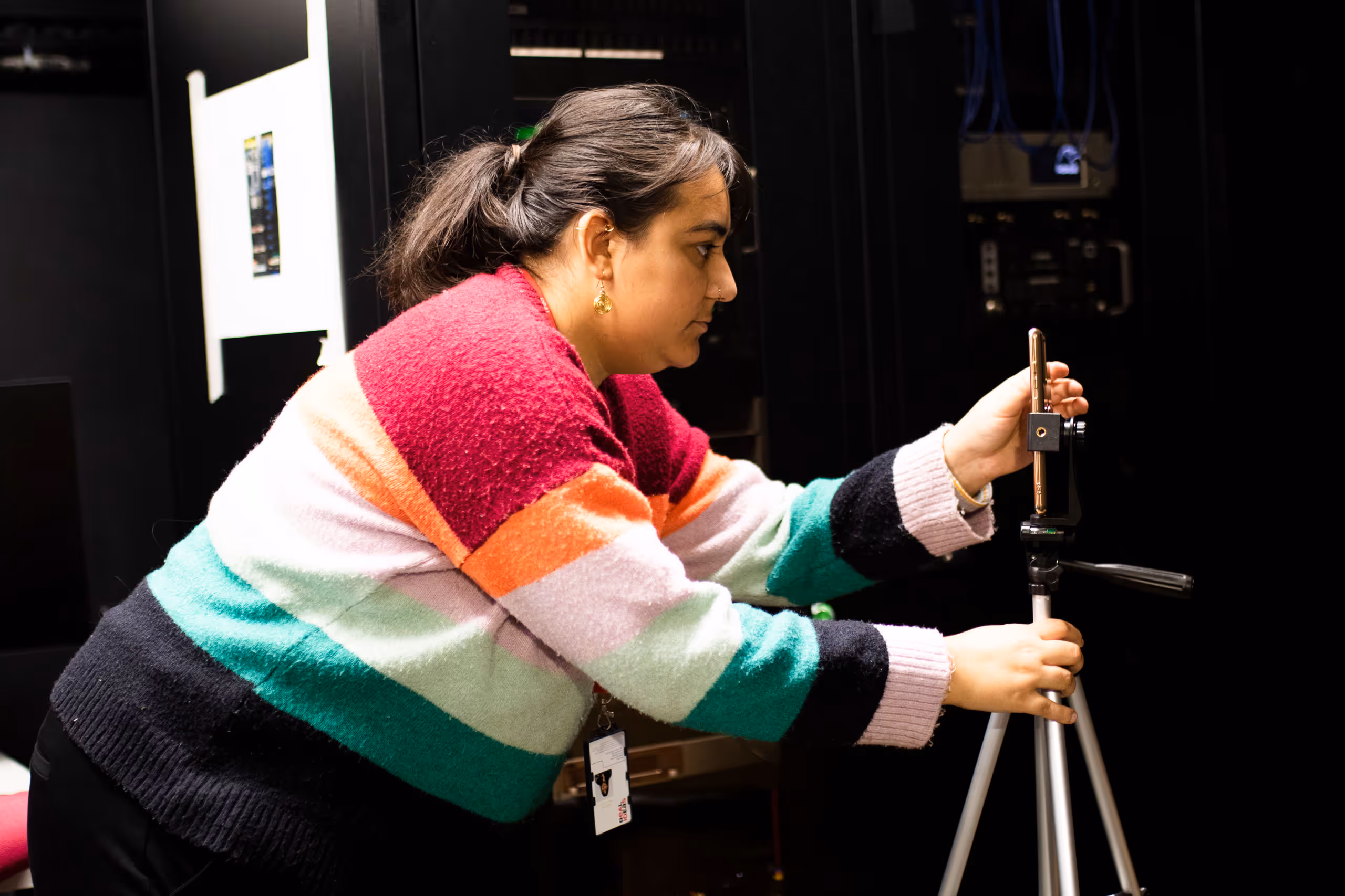 Young woman in colorful striped sweater adjusting camera equipment on tripod in dark venue