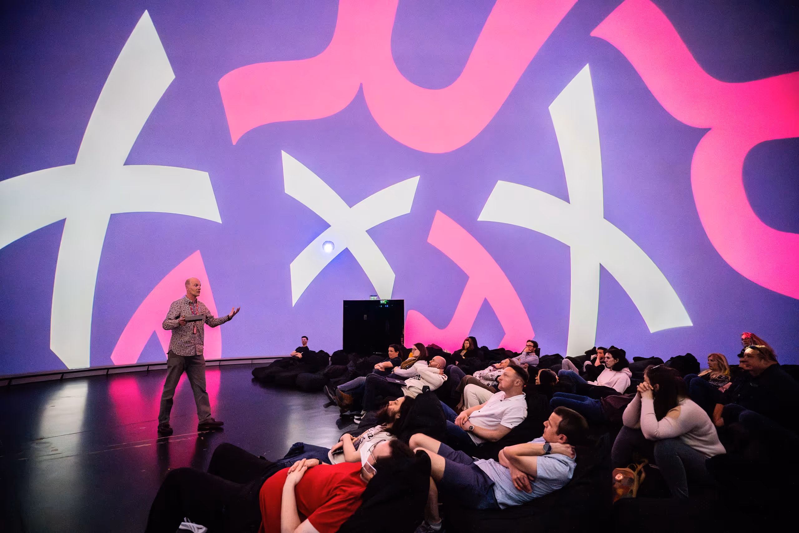 Speaker presenting to relaxed audience sitting on floor in front of large purple and pink graphic display