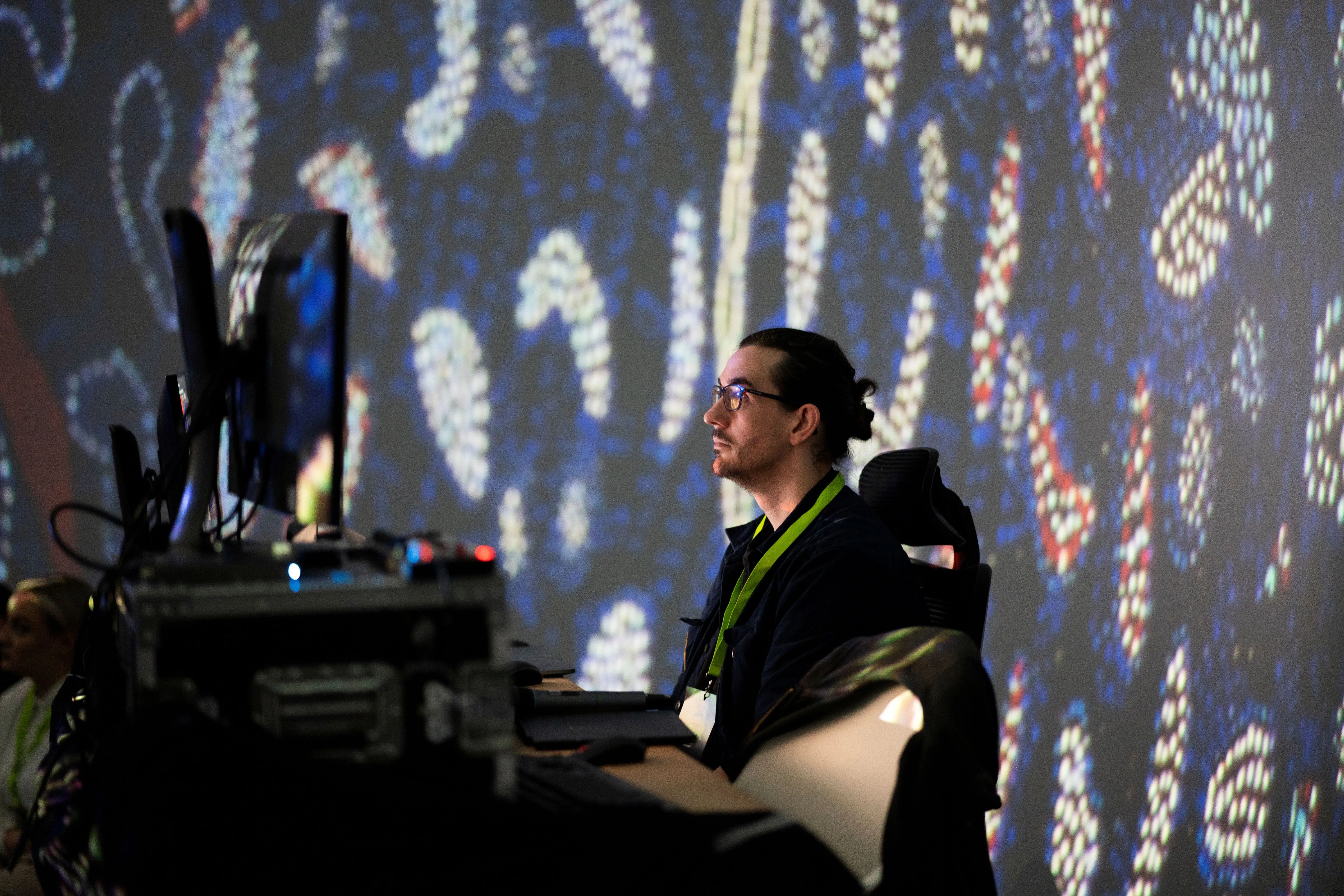 Man with glasses and a green lanyard sitting at a desk with multiple computer monitors, colorful abstract patterns projected on the wall behind him.