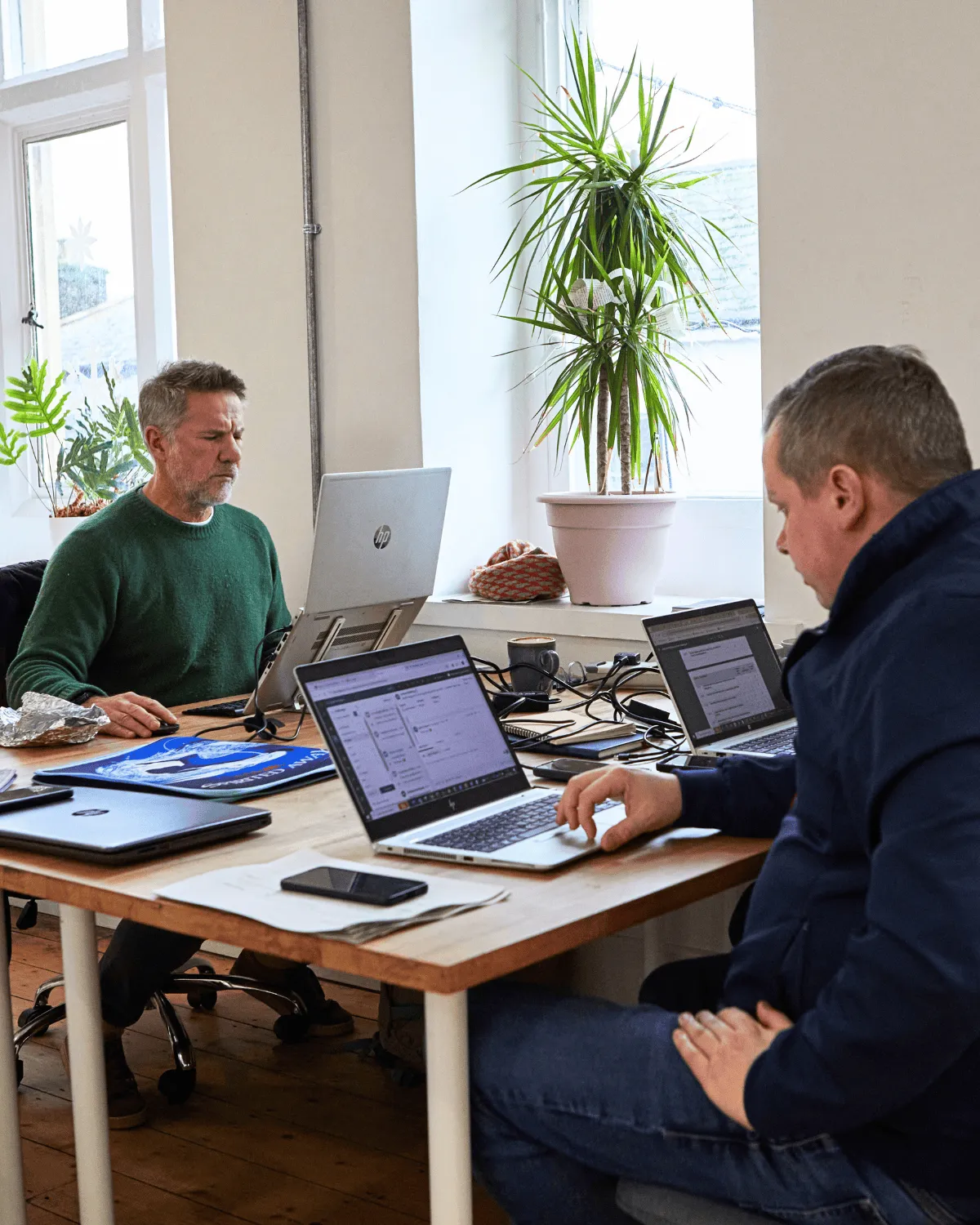 Two men working on laptops at a wooden table in a bright room with large windows and potted plants.