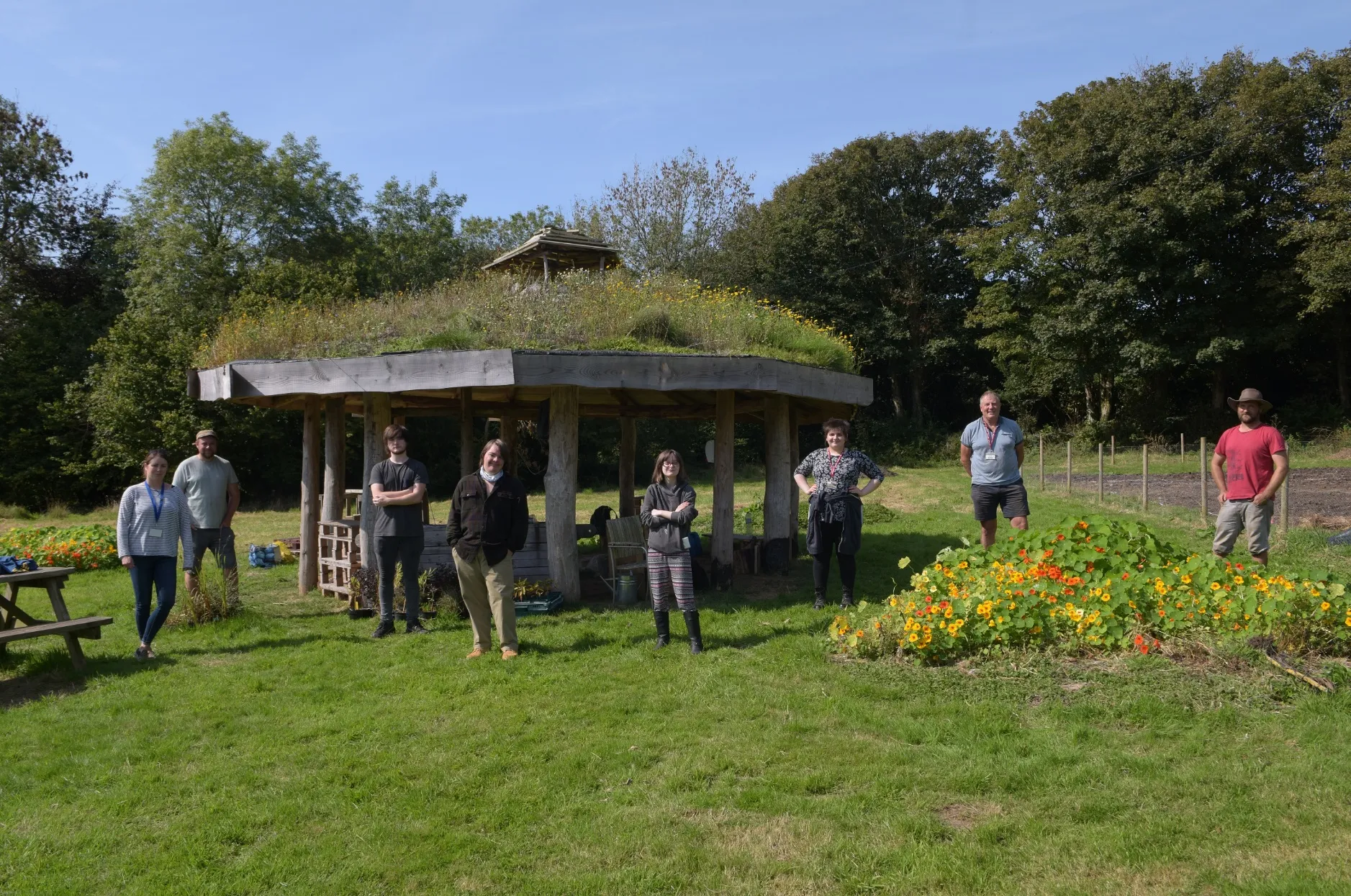 Group of seven people standing spaced apart on green grass in front of a wooden shelter with a grass roof, with trees and flowers in the background.
