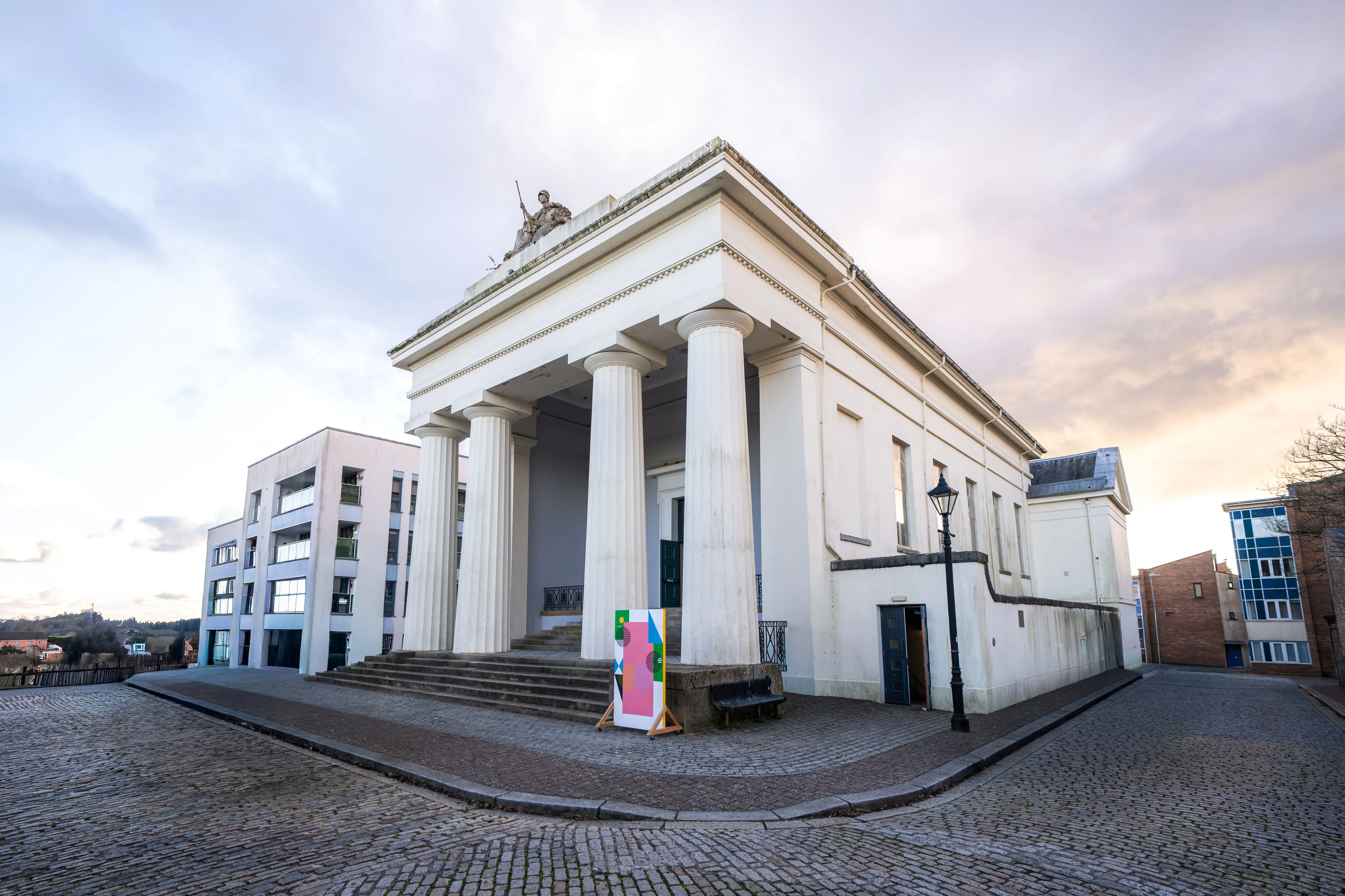 Neoclassical white building with large columns and statue on roof, located on a cobblestone street corner.