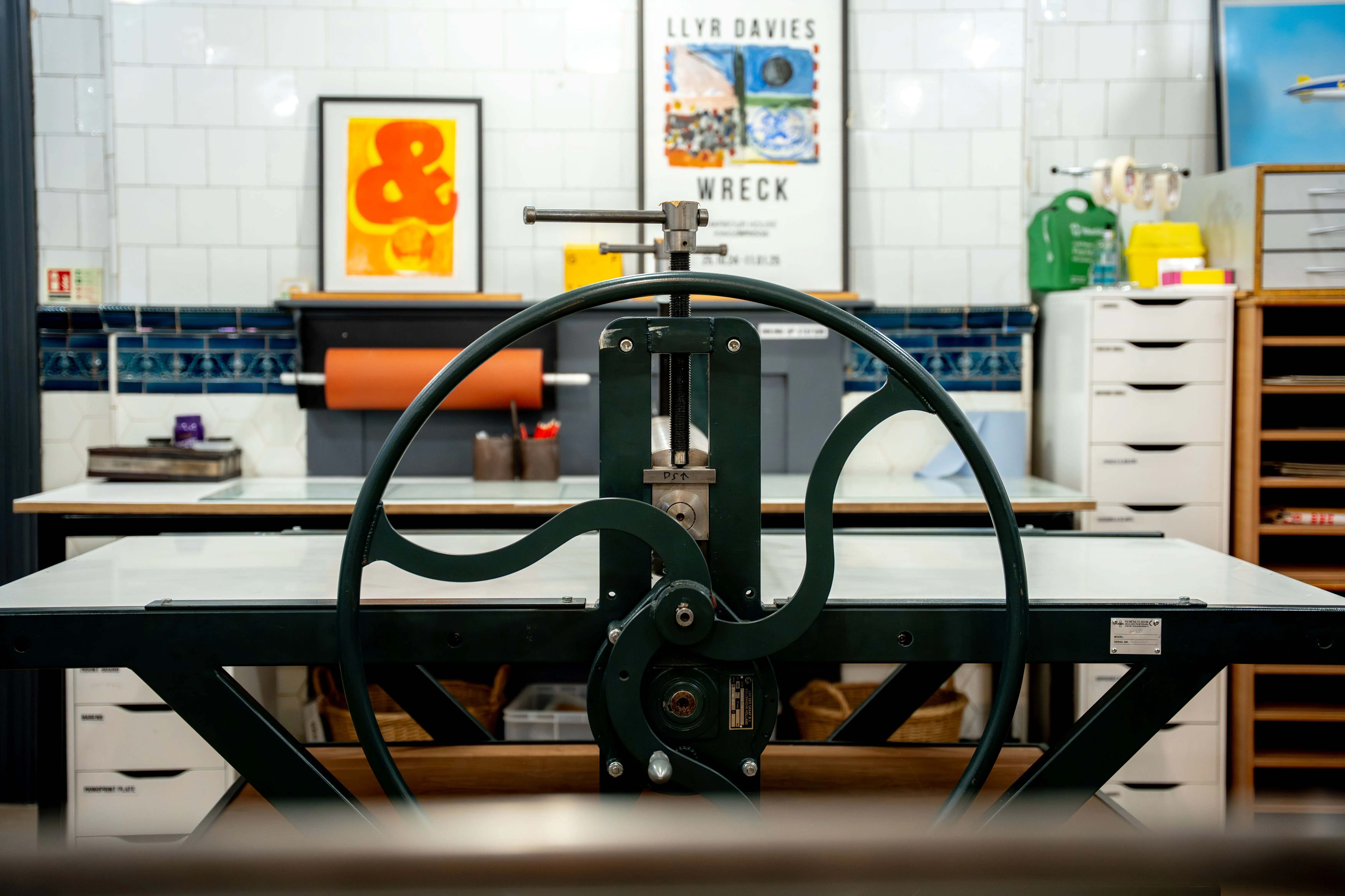 Close-up of a green printing press wheel in a bright art studio with framed prints and storage drawers in the background.