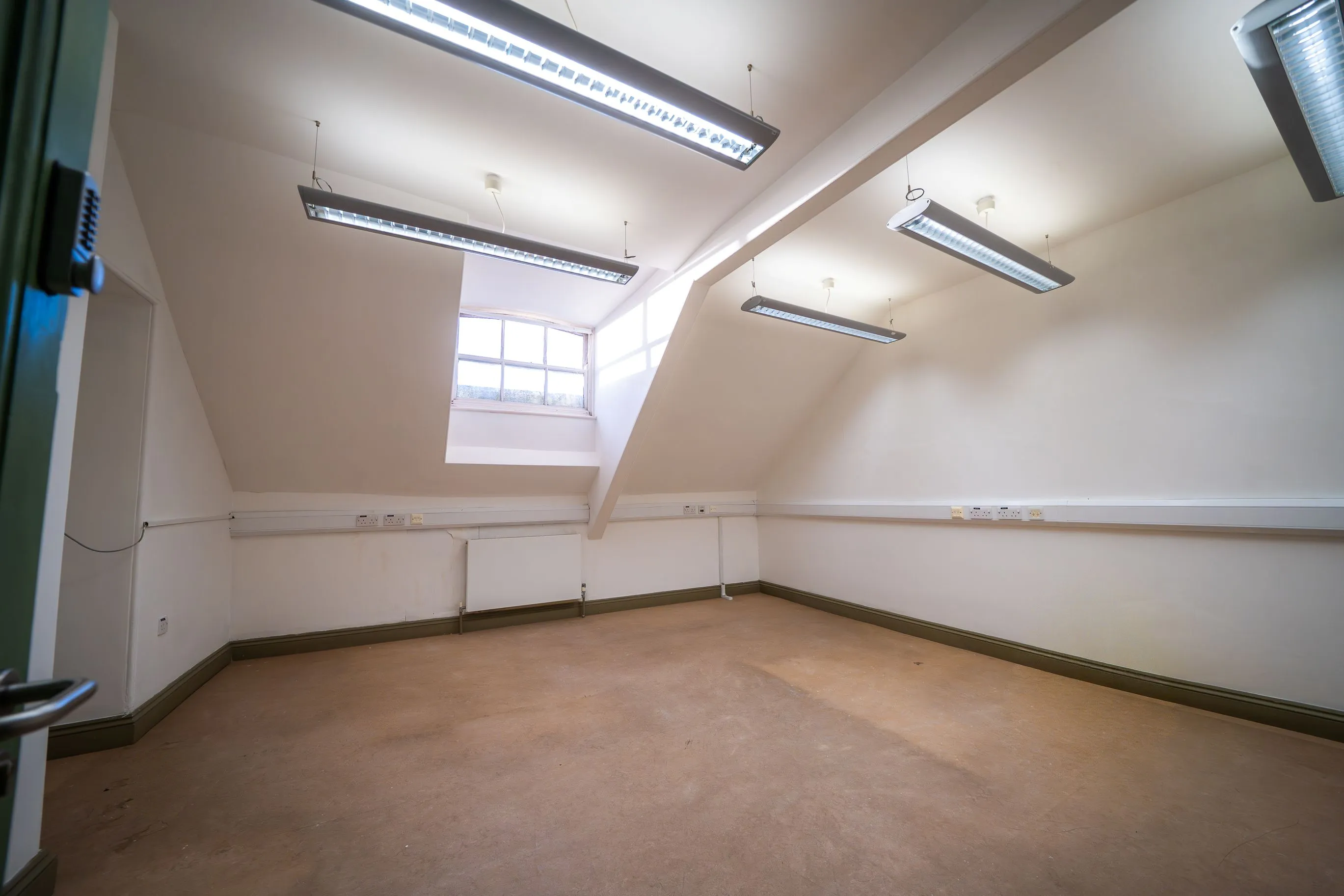 Empty room with beige floor, white walls, slanted ceiling, large window, and fluorescent ceiling lights.