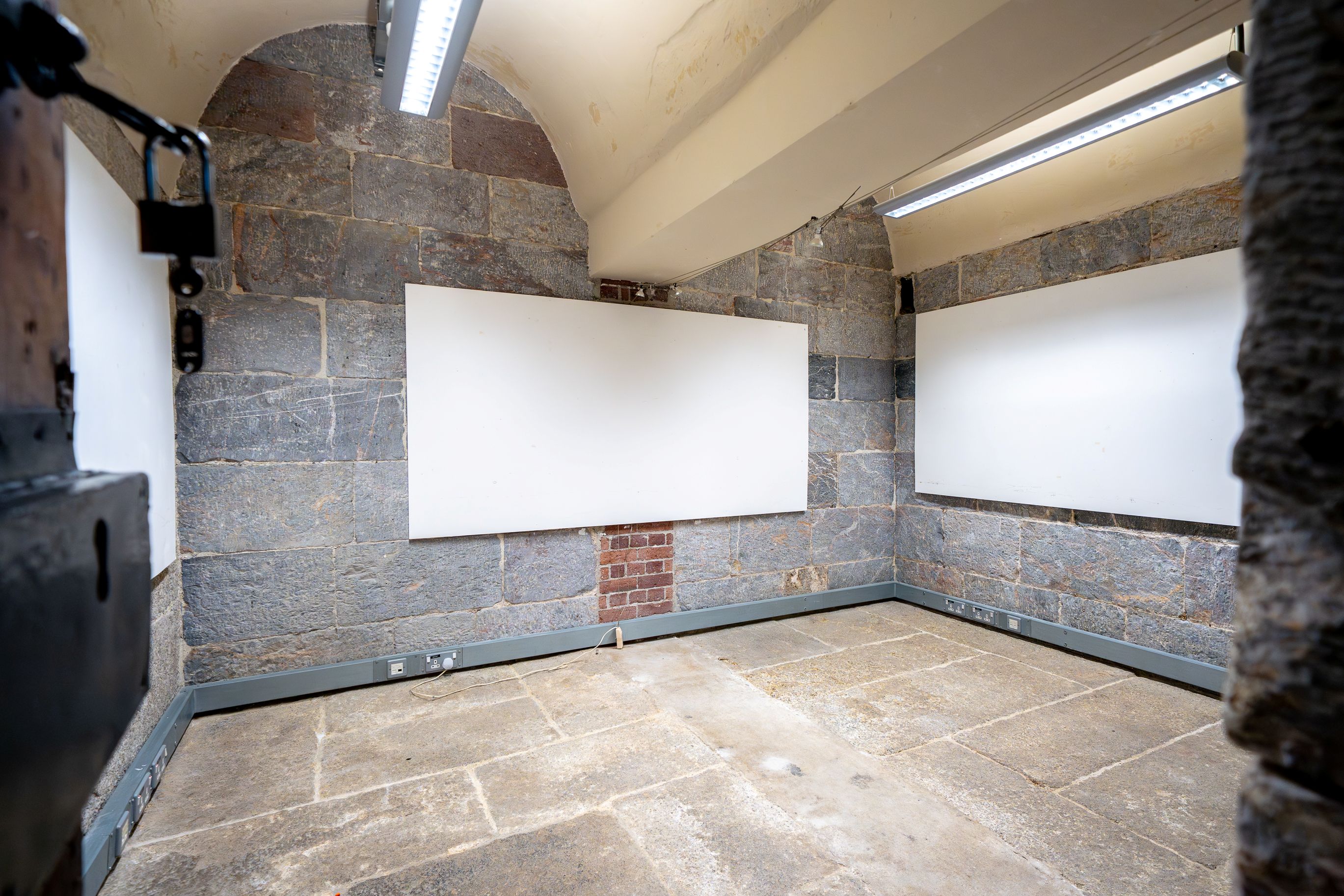 Empty stone-walled room with arched ceiling, two large blank whiteboards on the walls, and fluorescent lighting.
