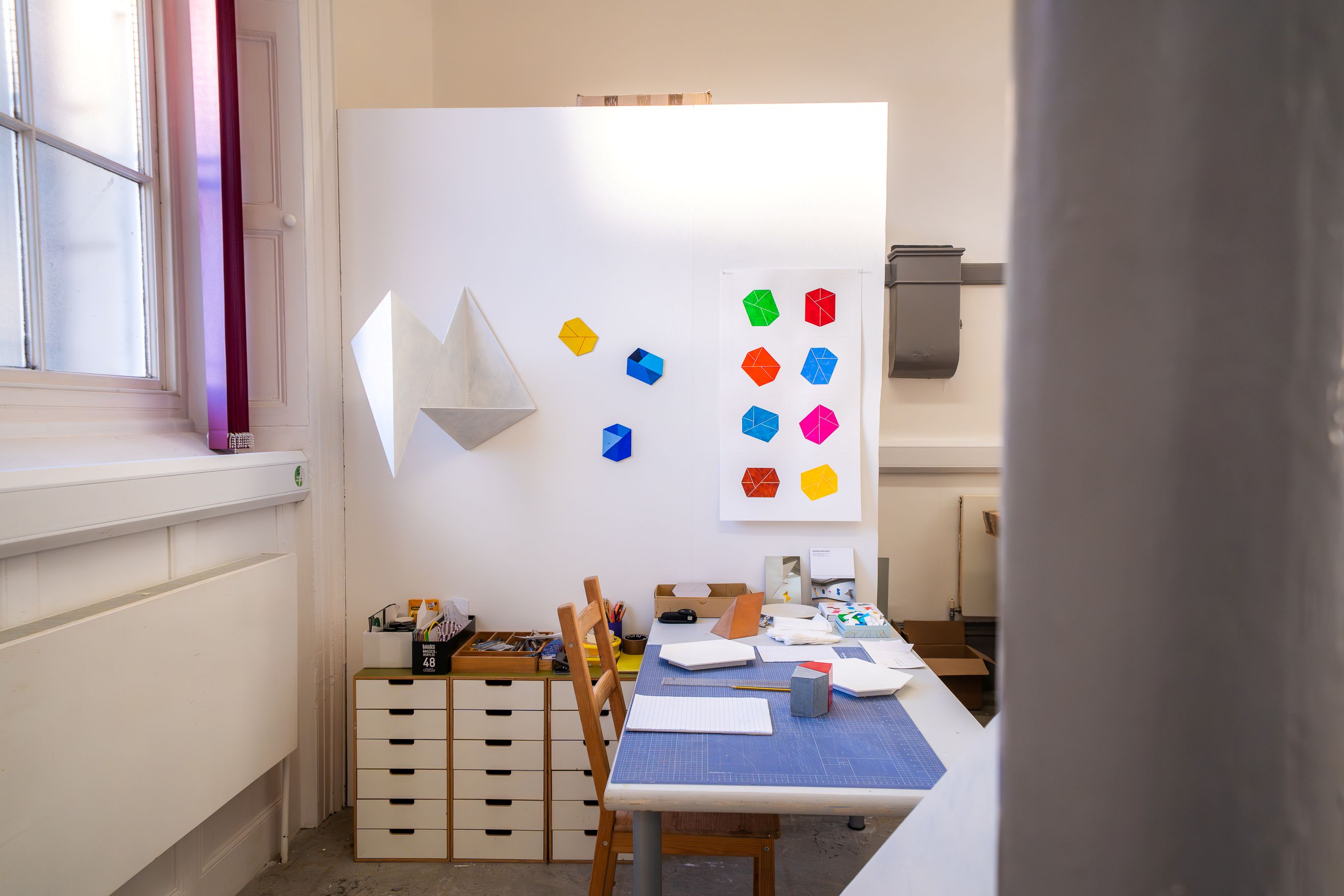 A bright art workspace with a white wall displaying colorful geometric shapes and folded paper models, a desk with cutting mats, papers, and art supplies, and two wooden chairs.