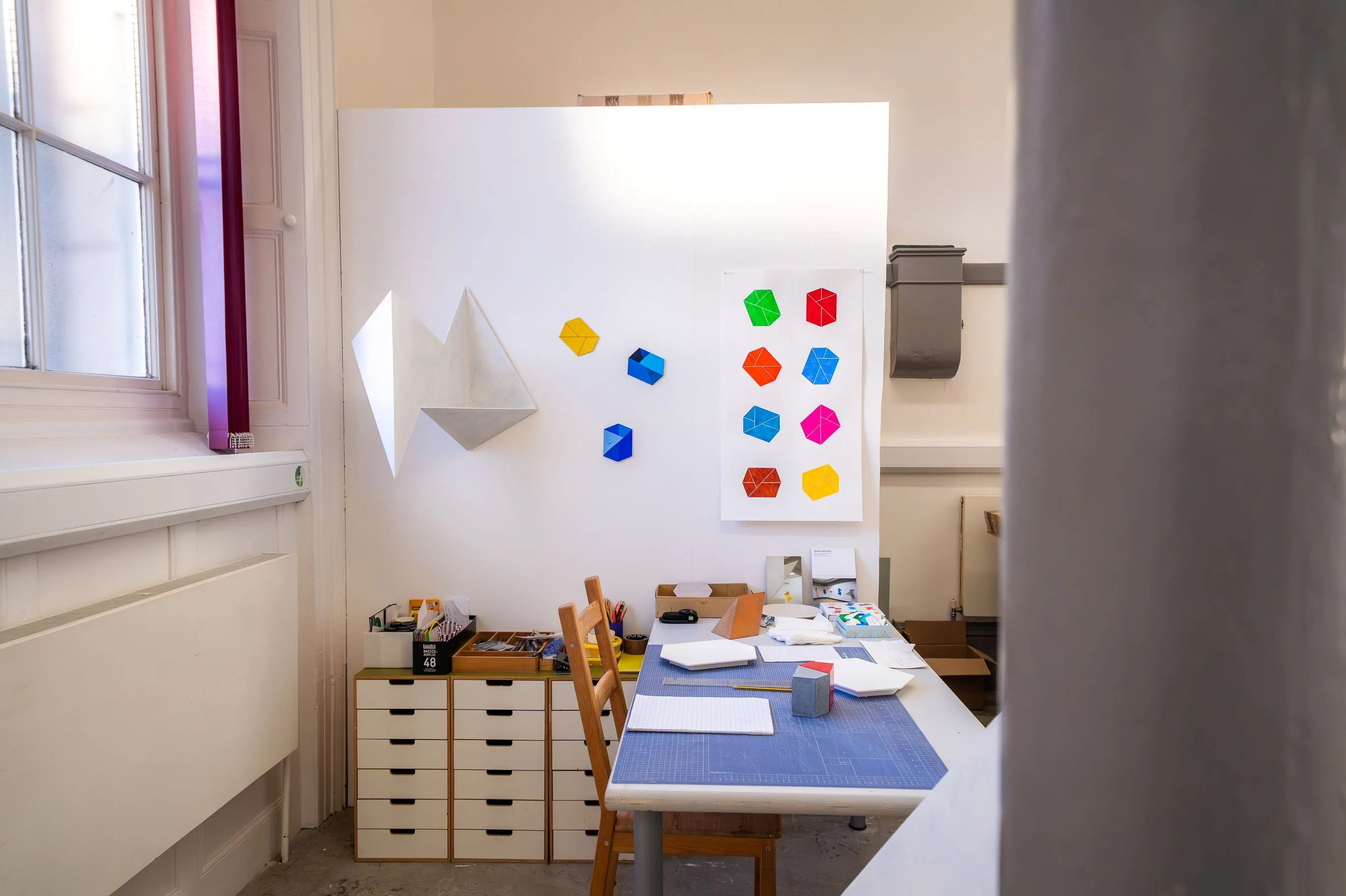 A bright art workspace with a white wall displaying colorful geometric shapes and folded paper models, a desk with cutting mats, papers, and art supplies, and two wooden chairs.