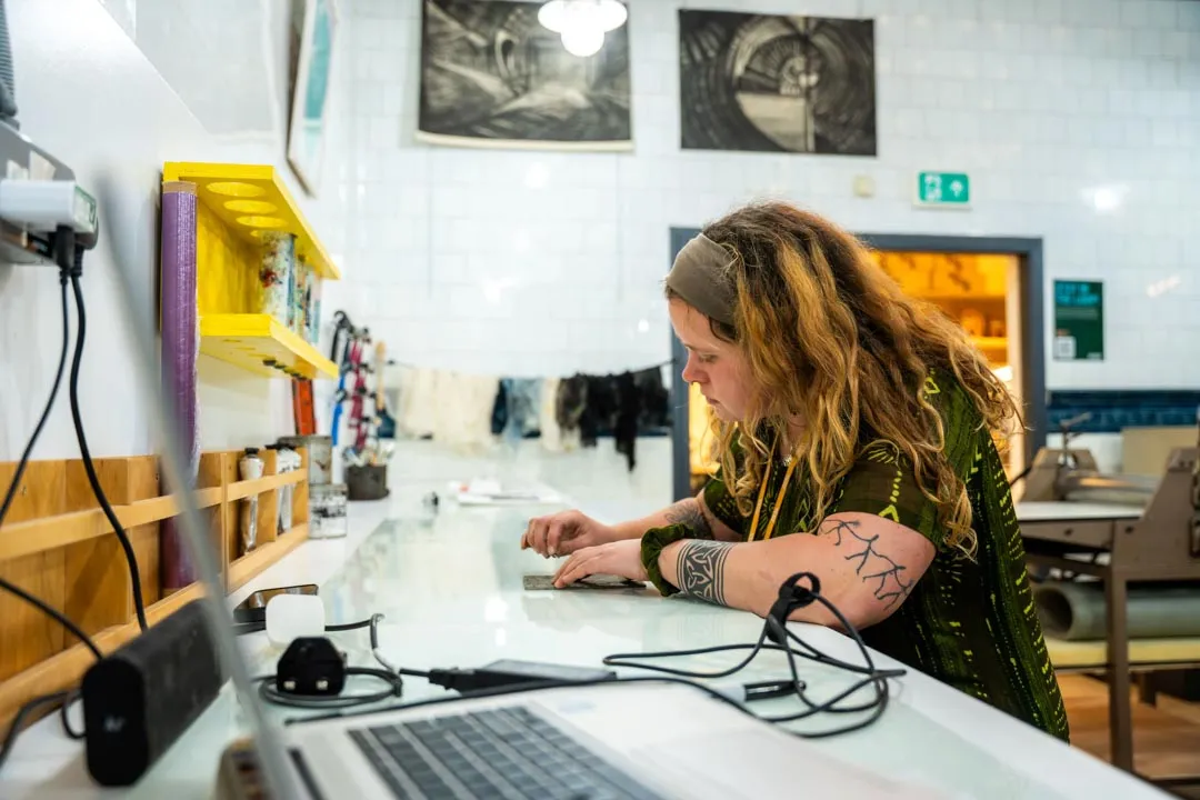 Person with long hair and tattoos focused on crafting or drawing on a worktable in an art studio.