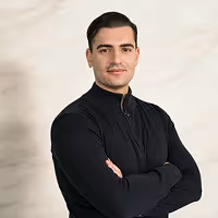 Junger Mann mit kurzen dunklen Haaren und Schnurrbart, gekleidet in schwarzes Oberteil, mit verschränkten Armen vor hellem Hintergrund.