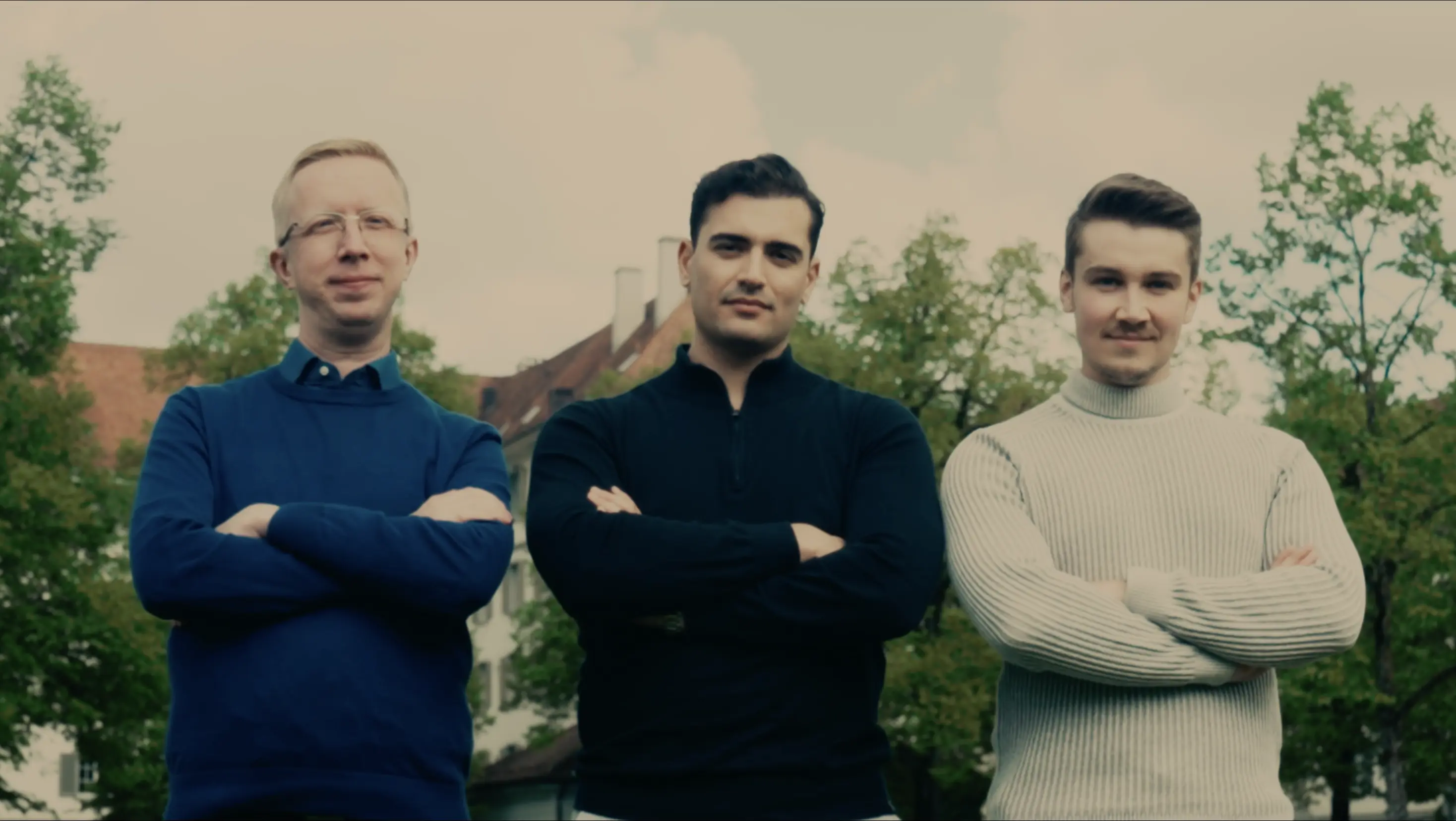 Drei Männer stehen mit verschränkten Armen und schauen in die Kamera, grüner Hintergrund mit Bäumen und Häusern.