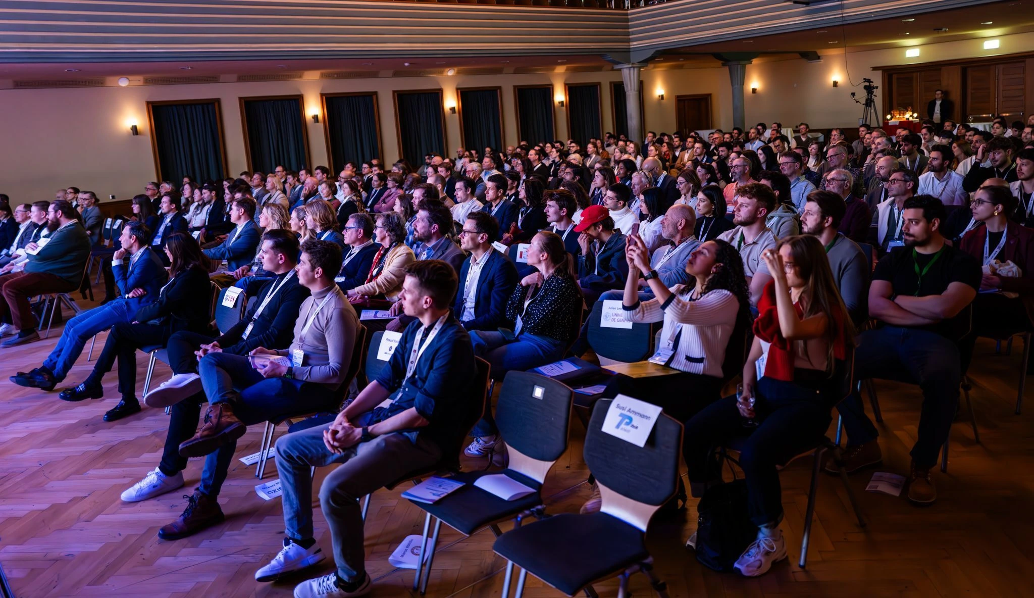 Ein vollbesetzter Veranstaltungssaal mit aufmerksam zuschauenden Personen bei einem Vortrag oder einer Konferenz.