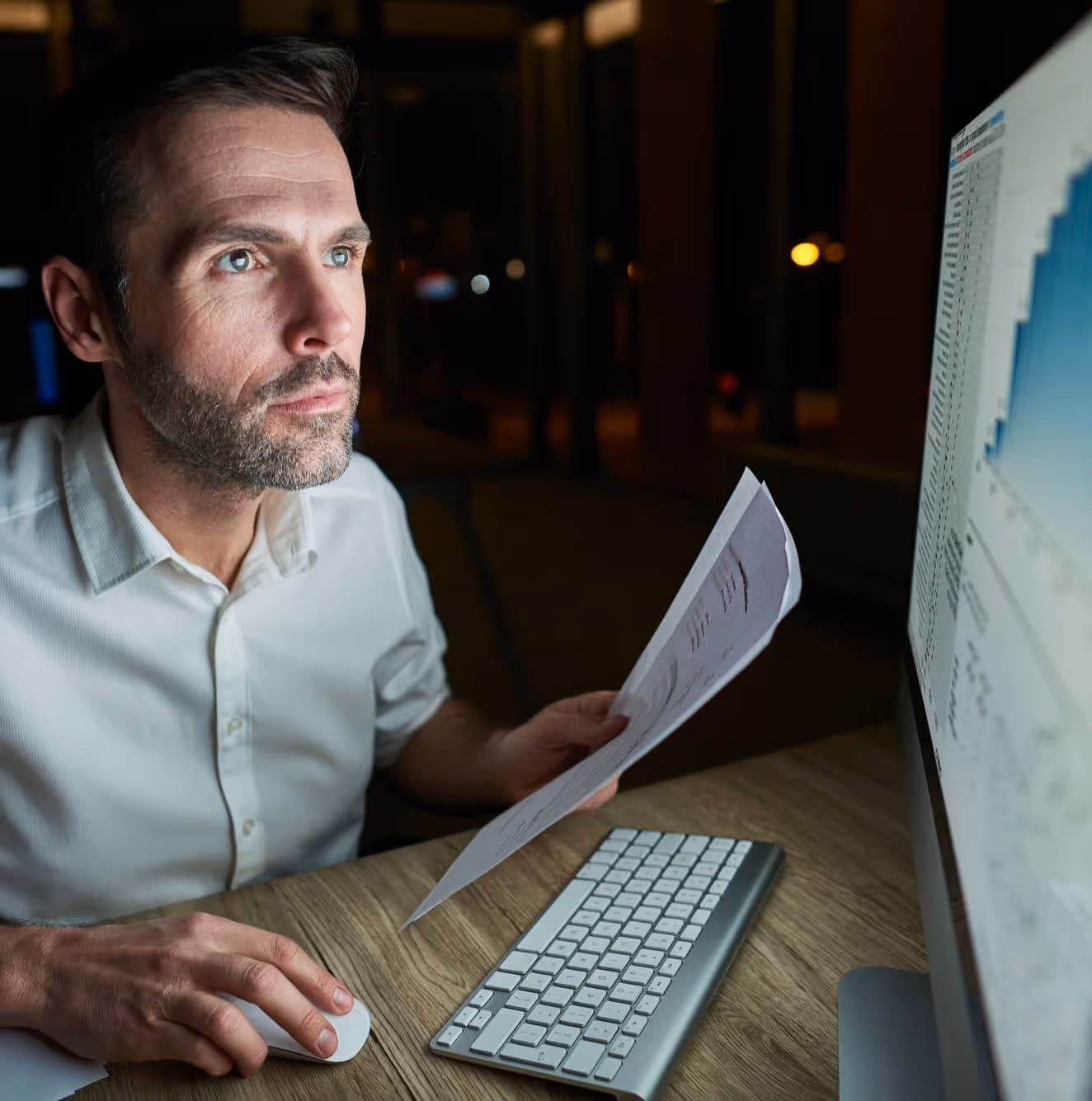Mann analysiert Daten auf Monitor und hält Dokumente, arbeitet spät am Schreibtisch.