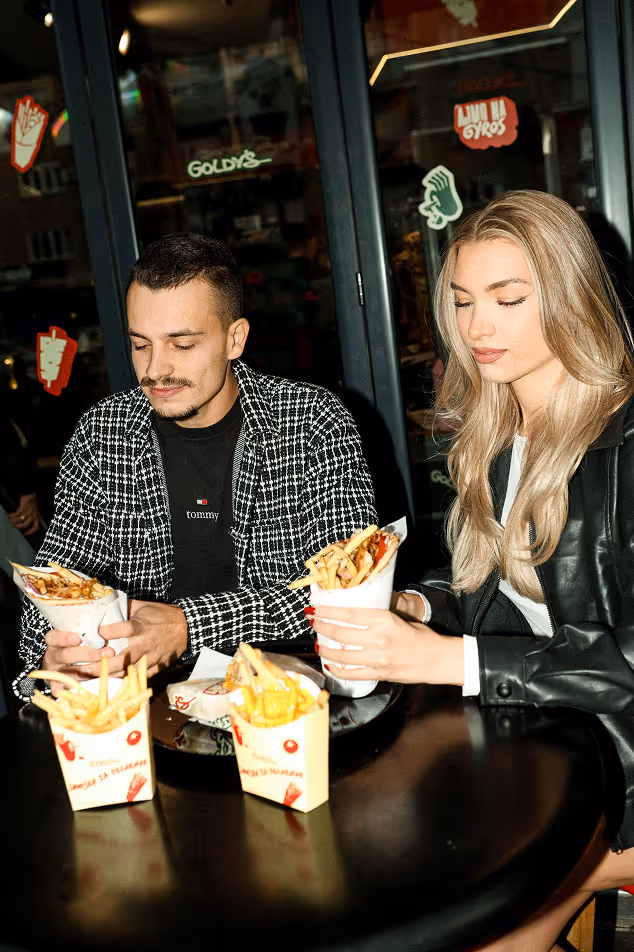Man and woman sitting at a table holding wrapped servings of fries or sandwiches inside a restaurant with dark glass doors and food stickers.