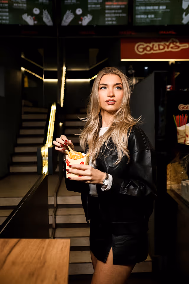 Young woman with long blonde hair holding a container of french fries inside a fast food restaurant.