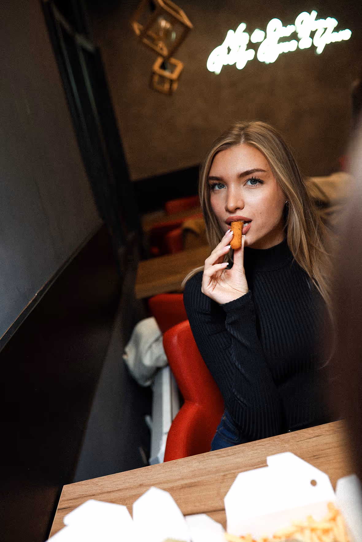 Plavokosa žena u crnom džemperu sedi u restoranu, jede prženu grickalicu i gleda u kameru.