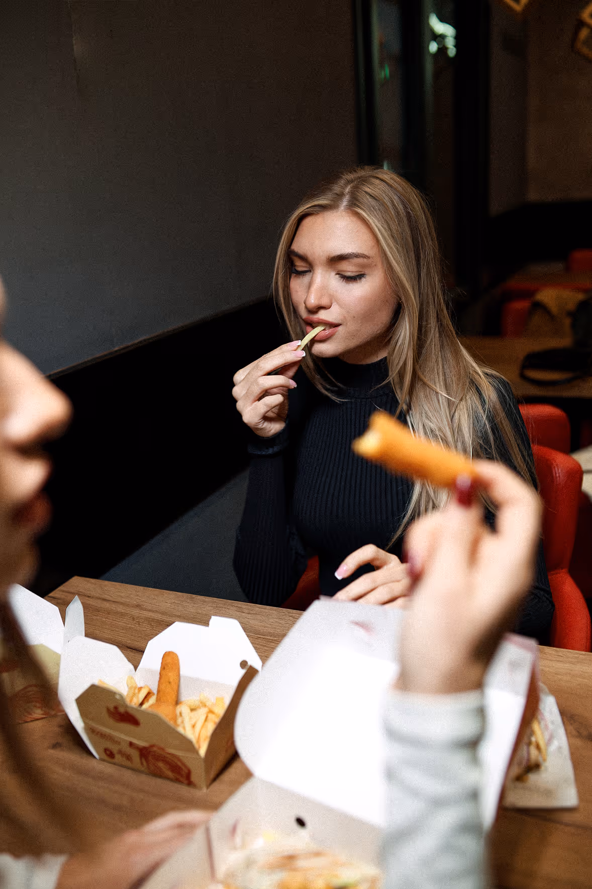 Mlada žena u crnom jede pomfrit za stolom sa pakovanjem hrane za poneti