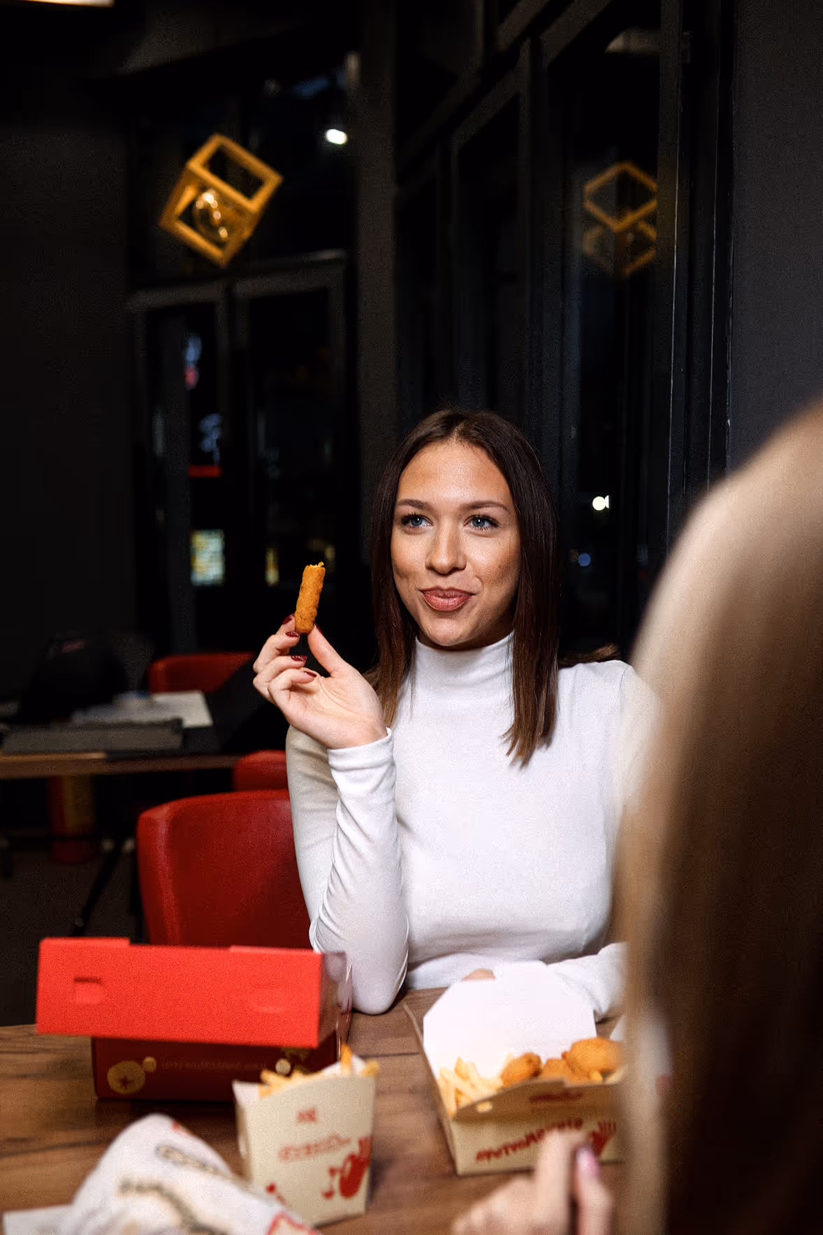 Žena u beloj rolki drži pileći nugget blizu usta, za stolom sa brzom hranom.