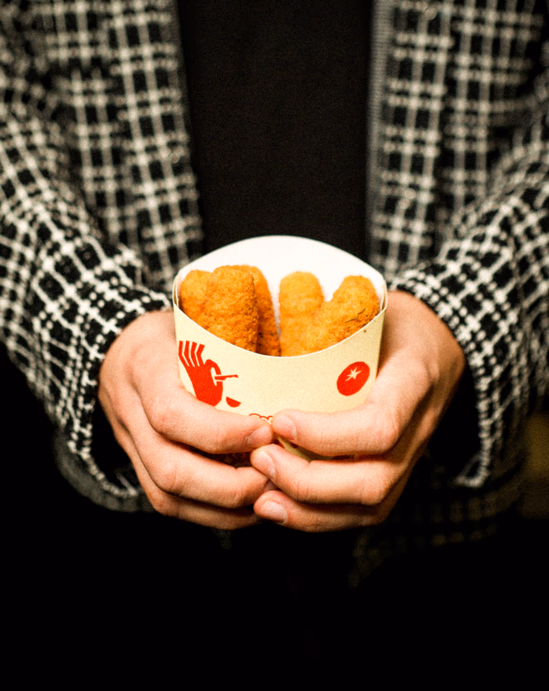 Person wearing a black and white checkered jacket holding a paper container with three breaded chicken nuggets.