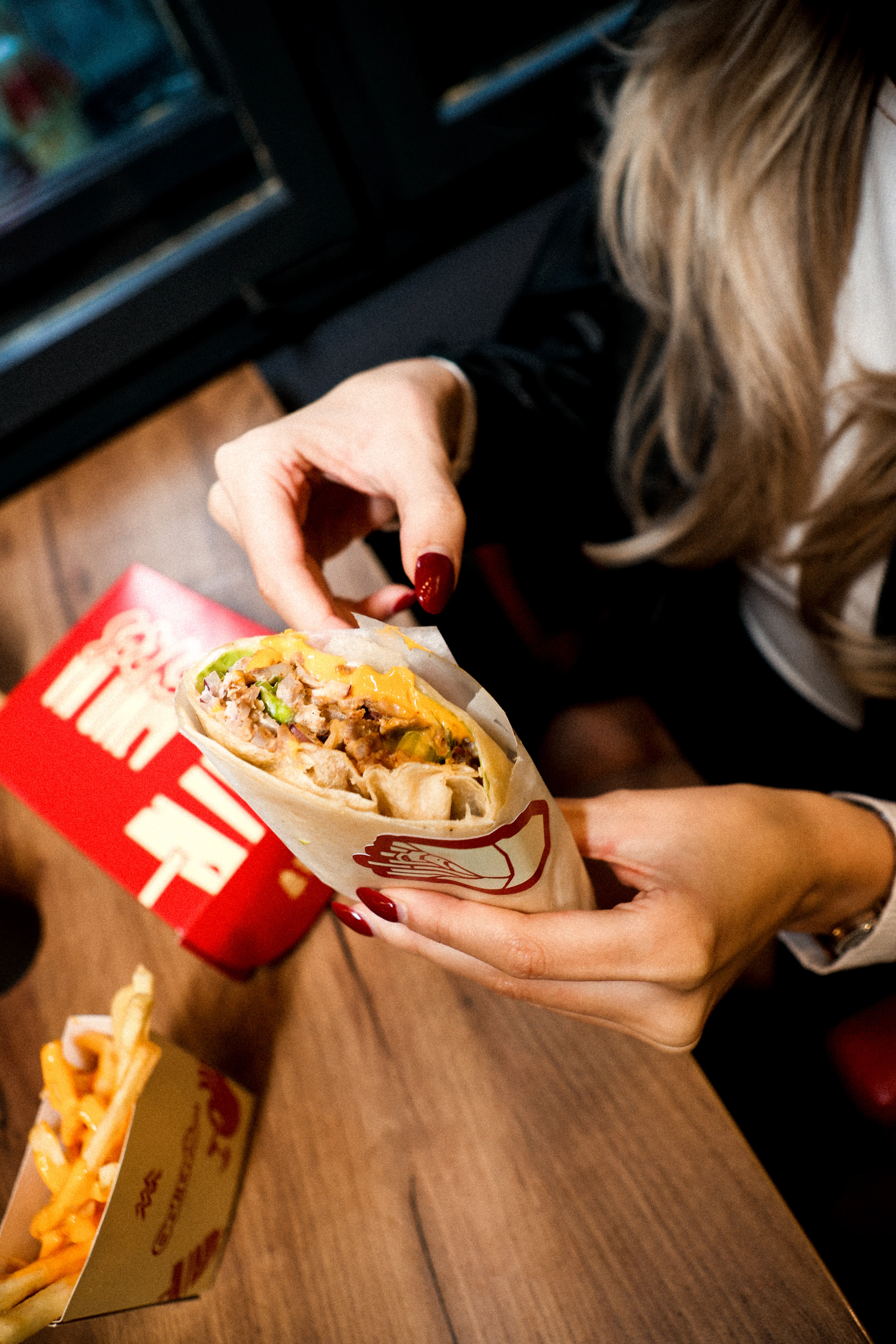 Osoba drži delimično odmotani burito sa pomfritom i crvenom kutijom za hranu na drvenom stolu.