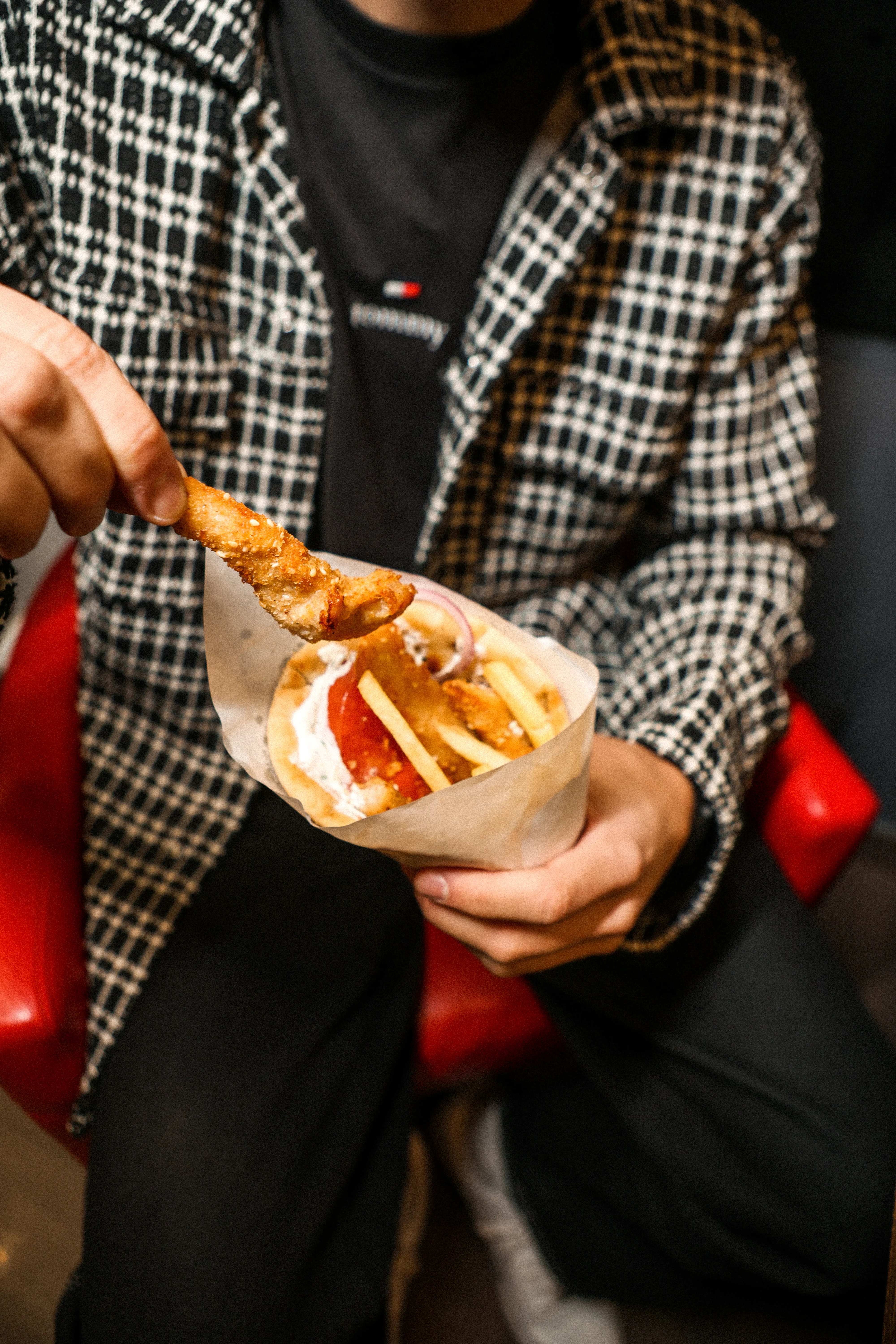 Osoba u crno-belom kariranom jaknom drži pitu punu pohovane piletine, pomfrita, paradajza, luka i sosa.