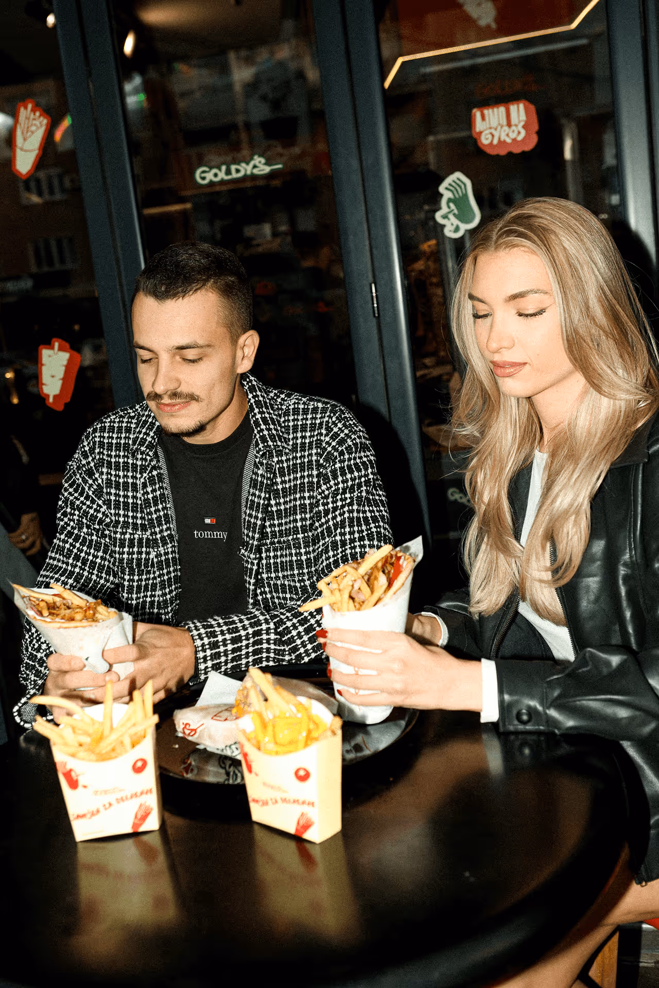Man and woman sitting at a table holding wrapped servings of fries or sandwiches inside a restaurant with dark glass doors and food stickers.