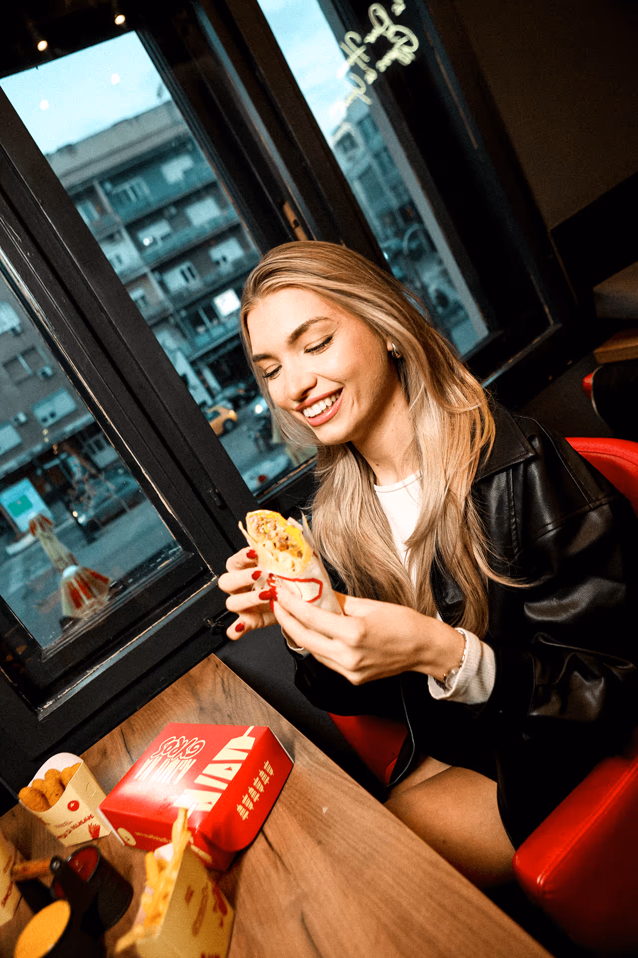 Smiling woman with long blonde hair sitting in a restaurant near a window, holding a wrapped sandwich.