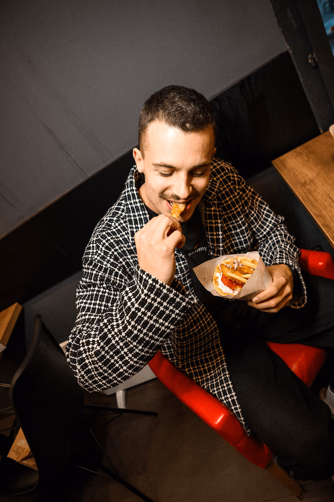 Smiling man in a black and white patterned jacket eating a sandwich with fries indoors.