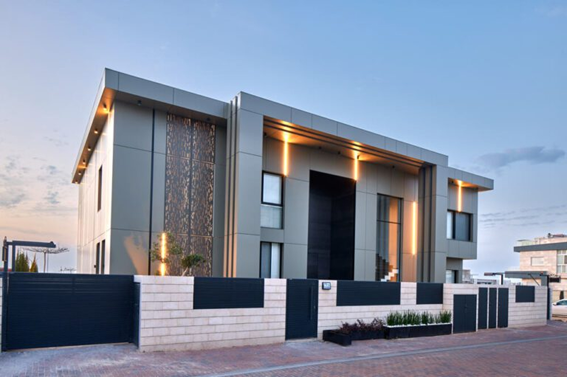 Modern two-story house with sleek gray panels, vertical accent lighting, and a black and beige brick fence in front.