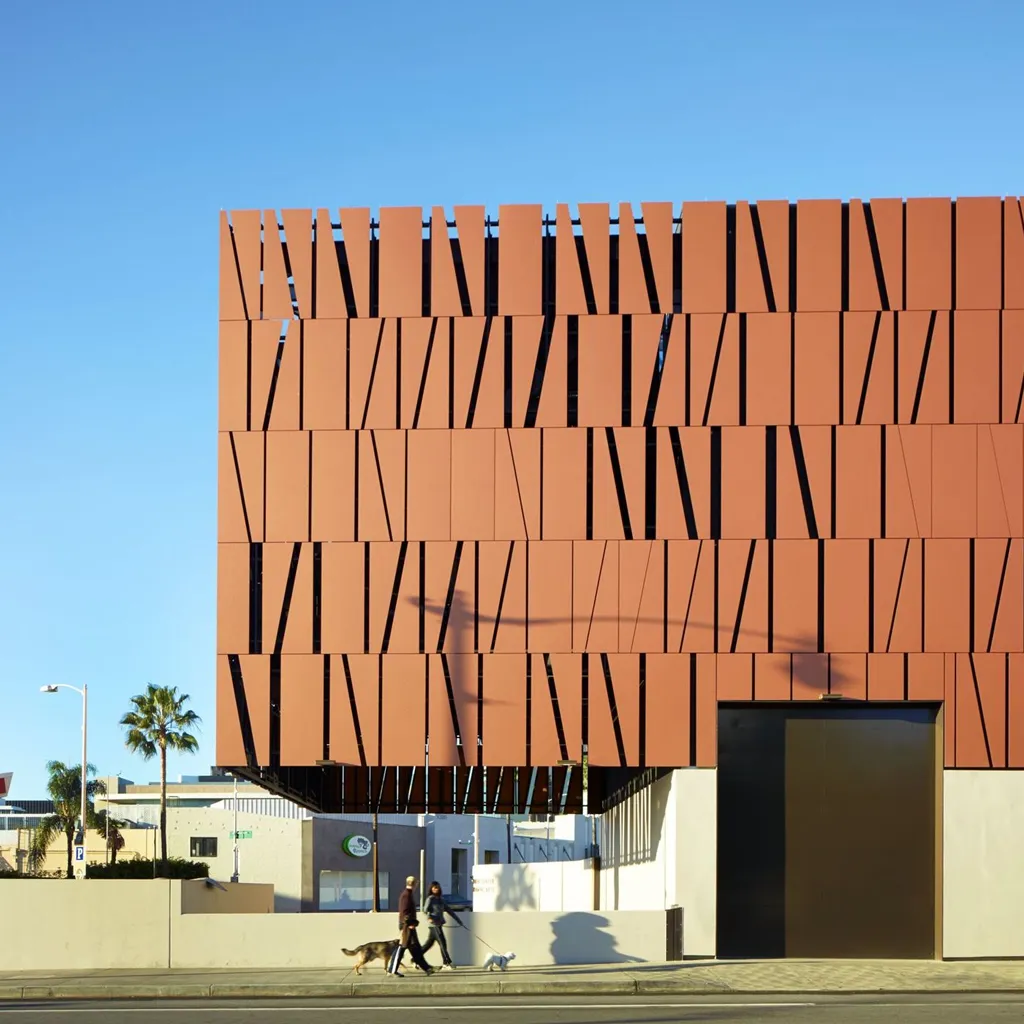 Two people walking dogs on sidewalk past a modern building with a geometric brown facade under a clear blue sky.