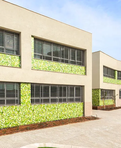 Modern beige building with large windows covered by horizontal blinds and green leafy decorative panels beneath the windows.