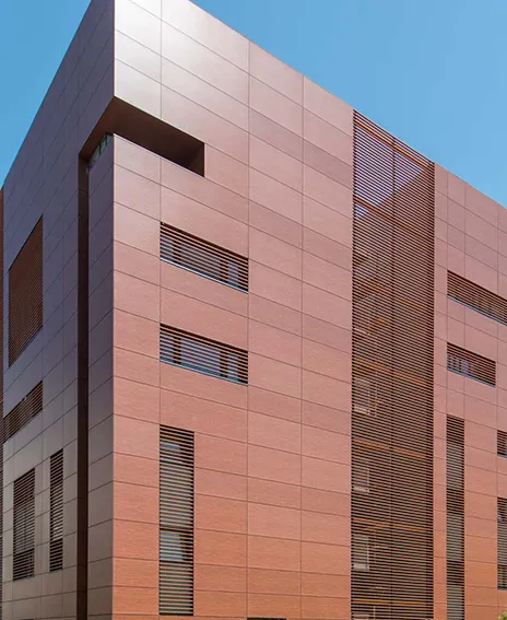 Modern building exterior with brown tiled facade featuring narrow horizontal and vertical windows with slatted blinds under a clear blue sky.