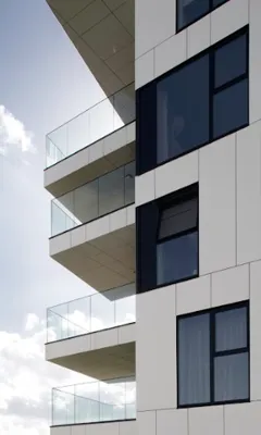Modern white building facade with glass balconies and square dark windows against a partly cloudy sky.