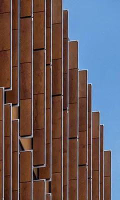 Close-up of a modern building facade with staggered vertical rust-colored metal panels against a clear blue sky.
