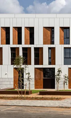 Modern building facade with white panels and tall vertical wooden window accents, surrounded by young trees.