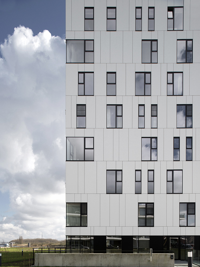 Modern building facade with a grid of rectangular windows reflecting cloudy sky and a grassy field beside it.