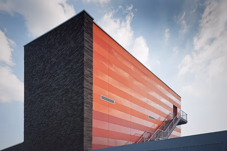 Modern building with dark brick facade on one side and red horizontal paneling on the other, featuring an external metal staircase under a partly cloudy sky.
