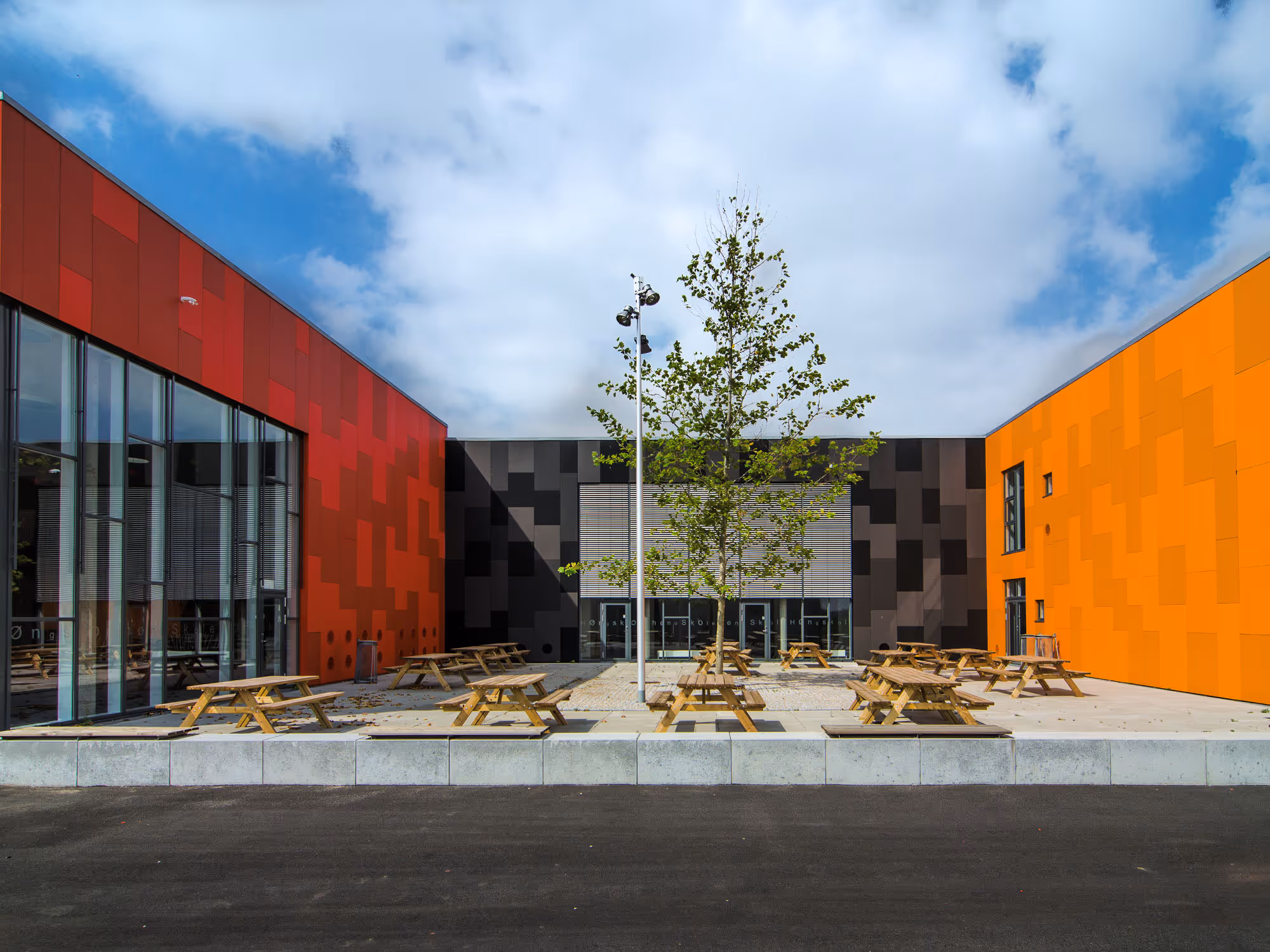 Courtyard with wooden picnic tables surrounded by modern buildings with red, black, and orange panel facades under a partly cloudy sky.