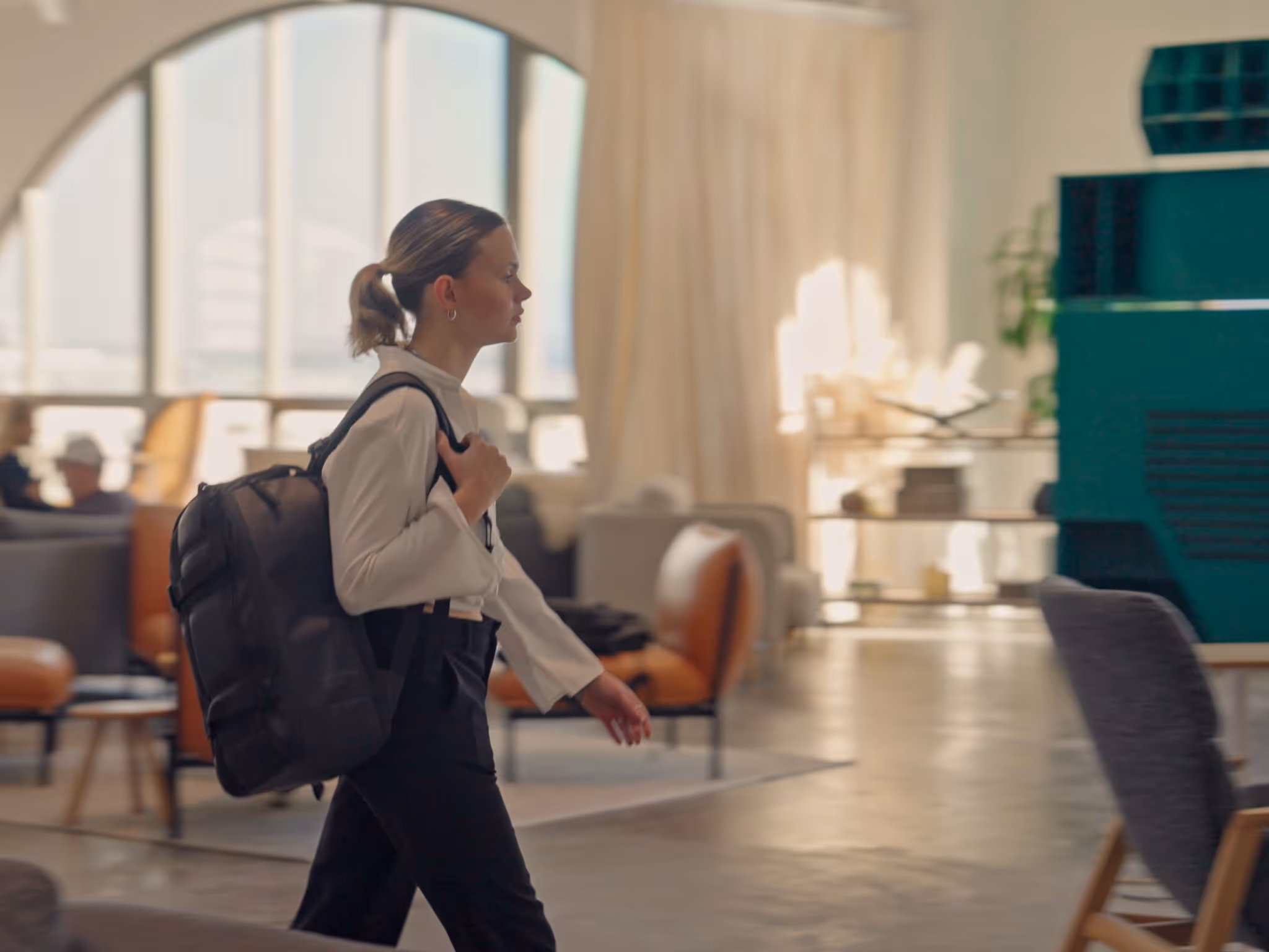 Woman with a ponytail wearing a white shirt and black pants walking indoors with a black backpack over one shoulder.