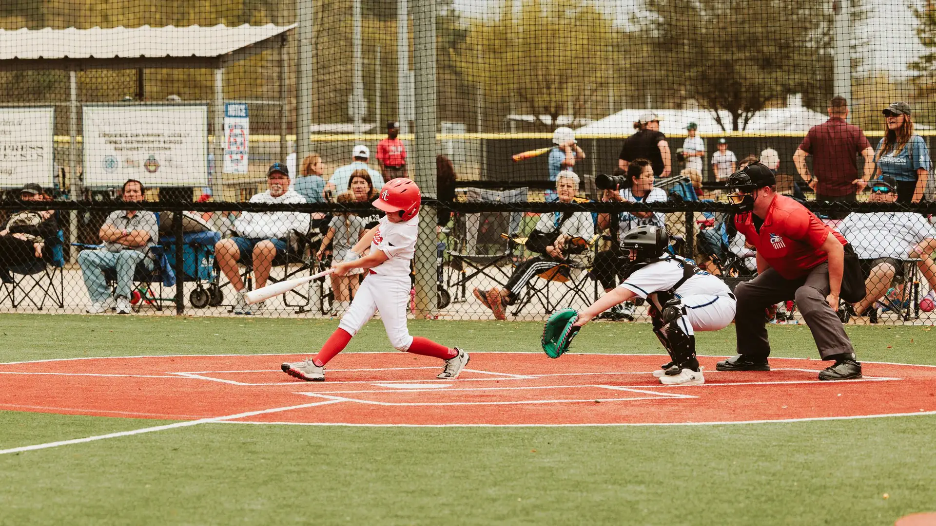 Young Chappapeela Sports Parks baseball player in red and white uniform swinging bat at home plate while catcher and umpire crouch behind, with spectators beyond the fence.