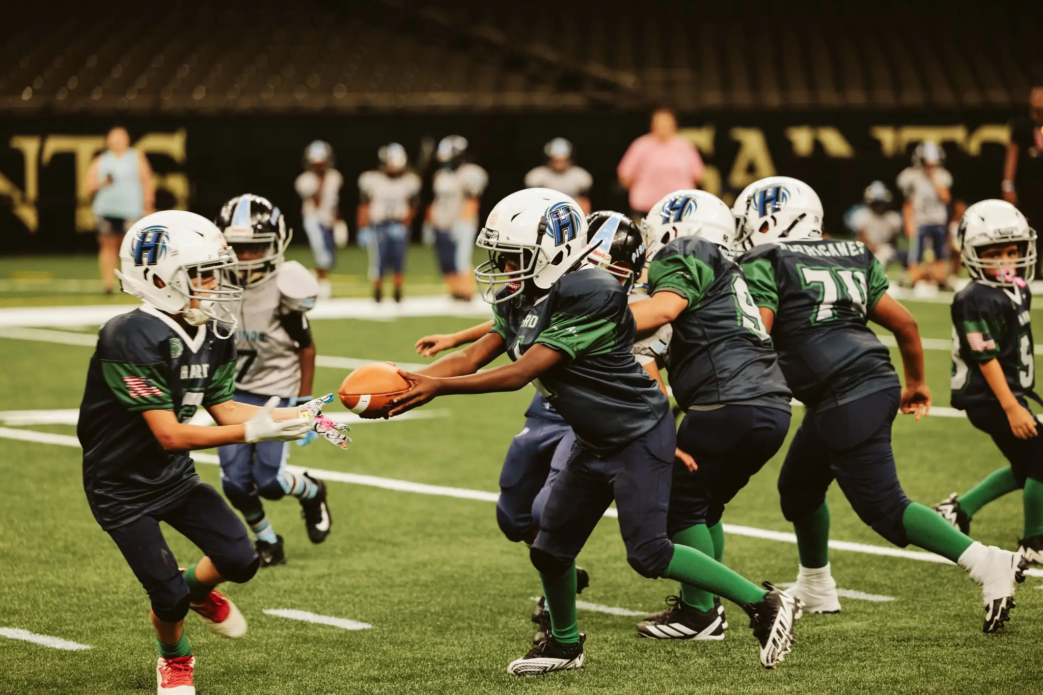 Youth H.A.R.D. #1 football players in dark green and navy uniforms with white helmets passing the ball on a football field.
